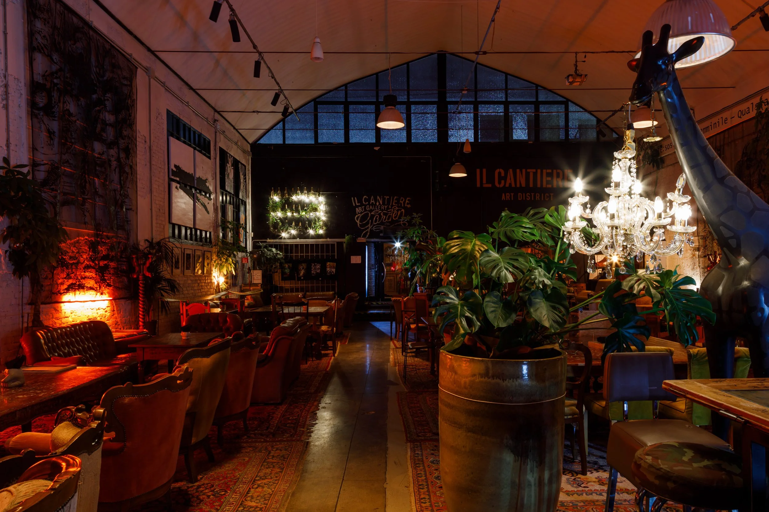 Interno di un bar o ristorante con arredamento vintage, luci calde, piante, sedie in velluto e un lampadario di cristallo, con decorazioni artistiche alle pareti e un grande vetro sul soffitto con vista notturna.