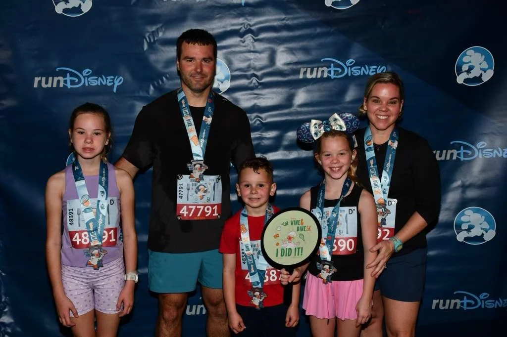 A family of five celebrating after completing a run Disney event, wearing medals and race bibs, standing in front of a blue backdrop with run Disney logos.