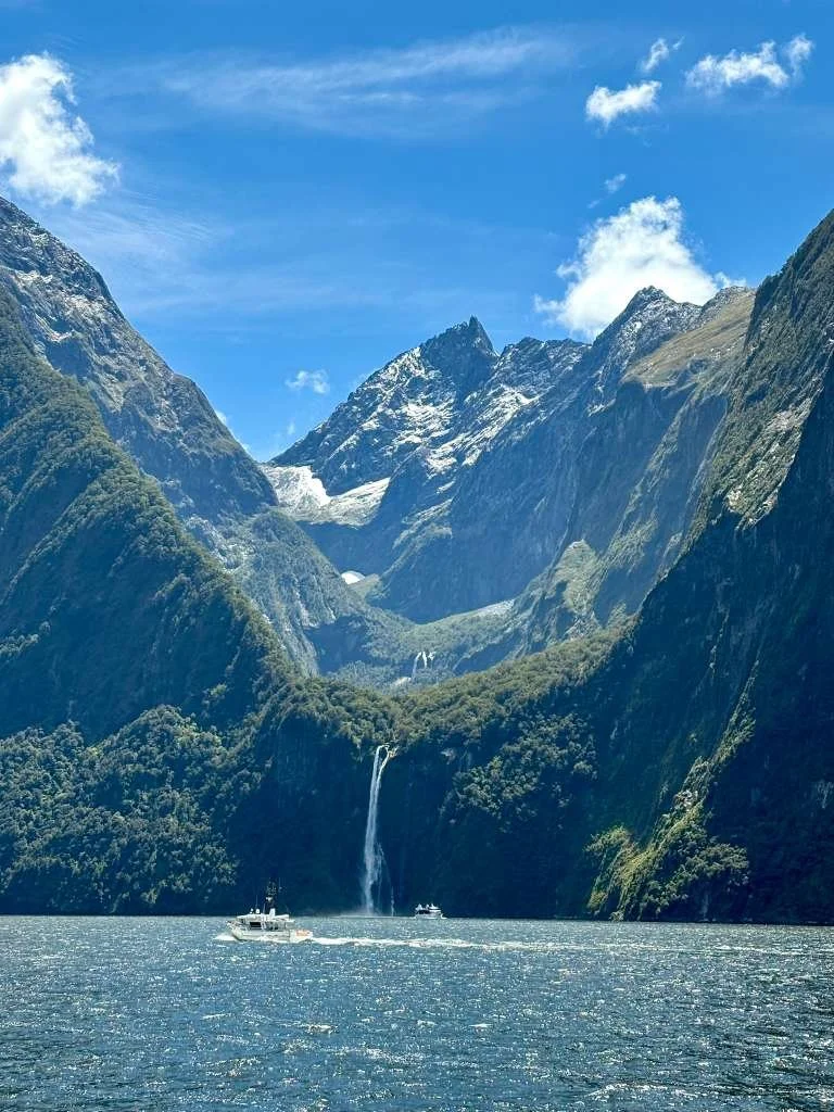 Scenic view of a fjord with boats in the water, tall mountains with snow-capped peaks in the background, a waterfall cascading into the fjord, and clear blue sky with some clouds.