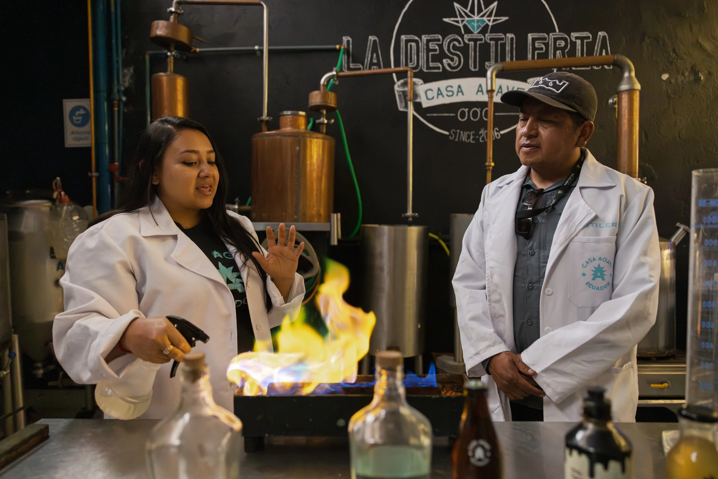 Two people in white lab coats working in a laboratory with chemical equipment. A fiery flame is burning in front of them, and there is a black wall with a logo that reads "La Destilerta Casa Aave" in the background.