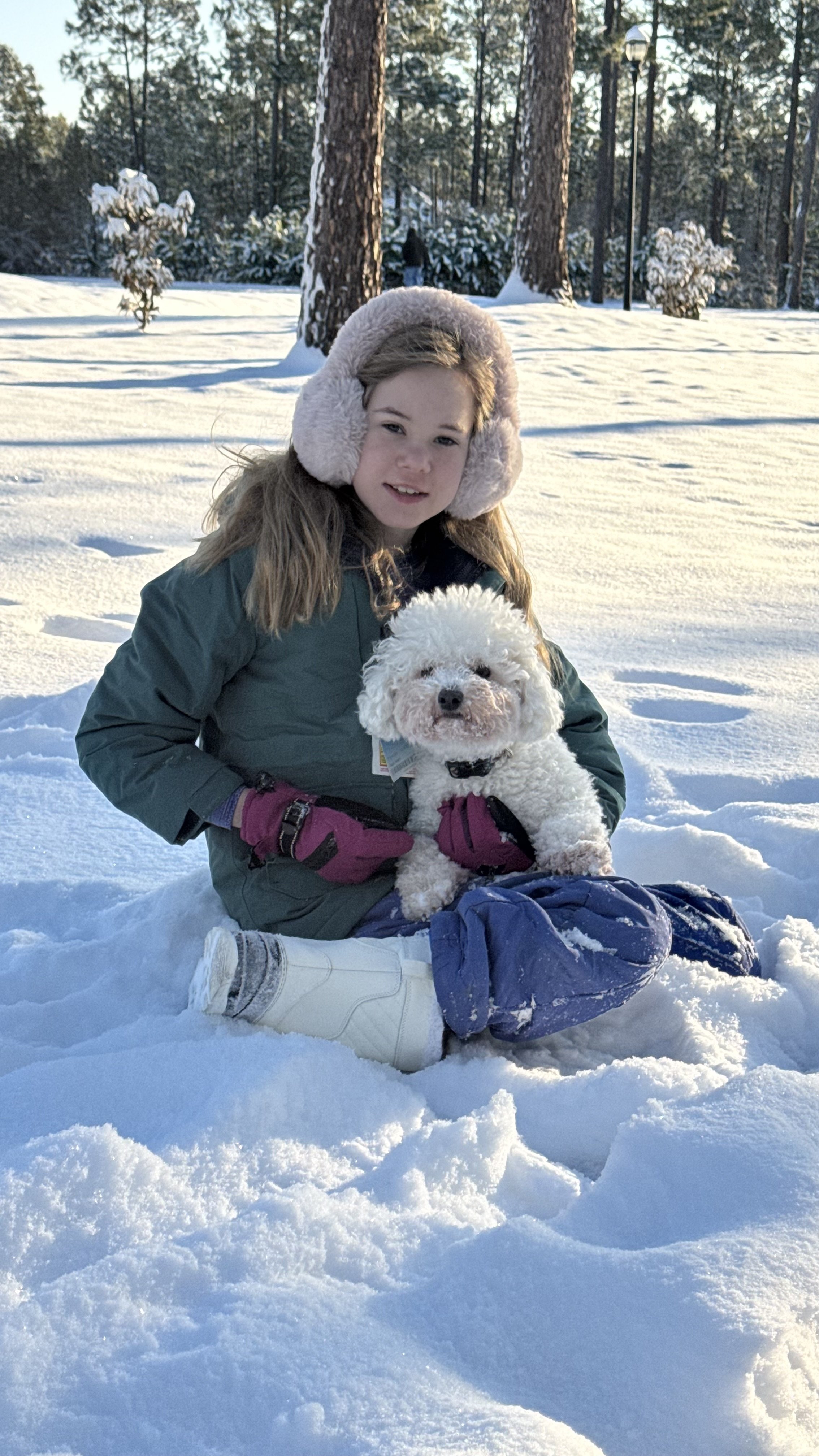 A girl wearing a jacket, ear muffs, gloves and snow boots holding a bichon frise  in the snow 