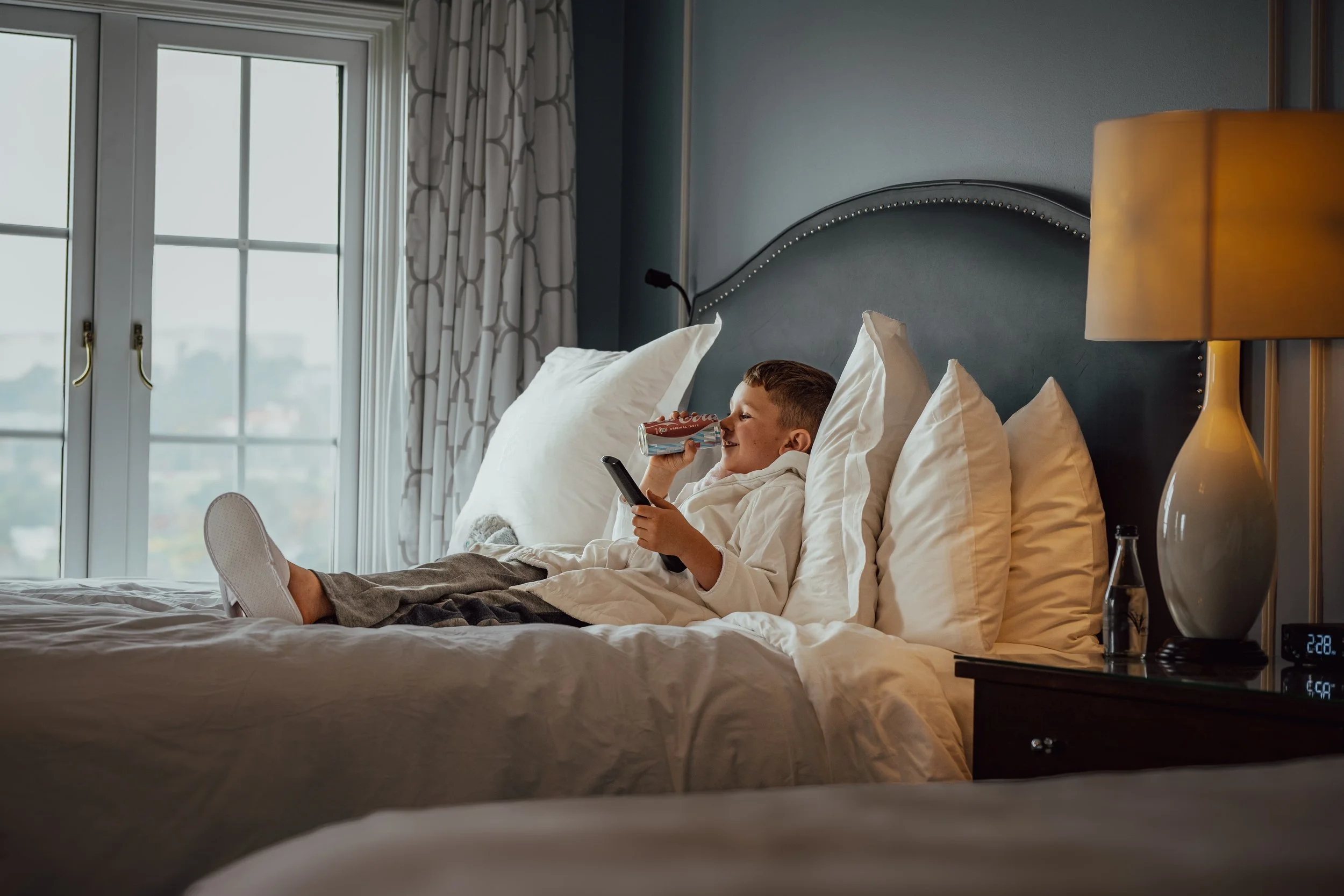 kid in bathrobe, in a plush bed with lots of pillows, window curtains drawn back, wearing slippers, with a TV remote in hand, and drinking Coca Cola at Four Seasons, the Westcliff, Johannesburg, South Africa