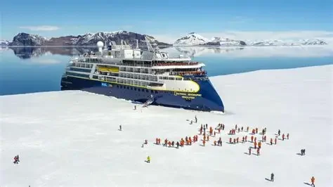 A large cruise ship docked in a snowy icy landscape with people in red jackets walking and gathering nearby under clear blue skies.
