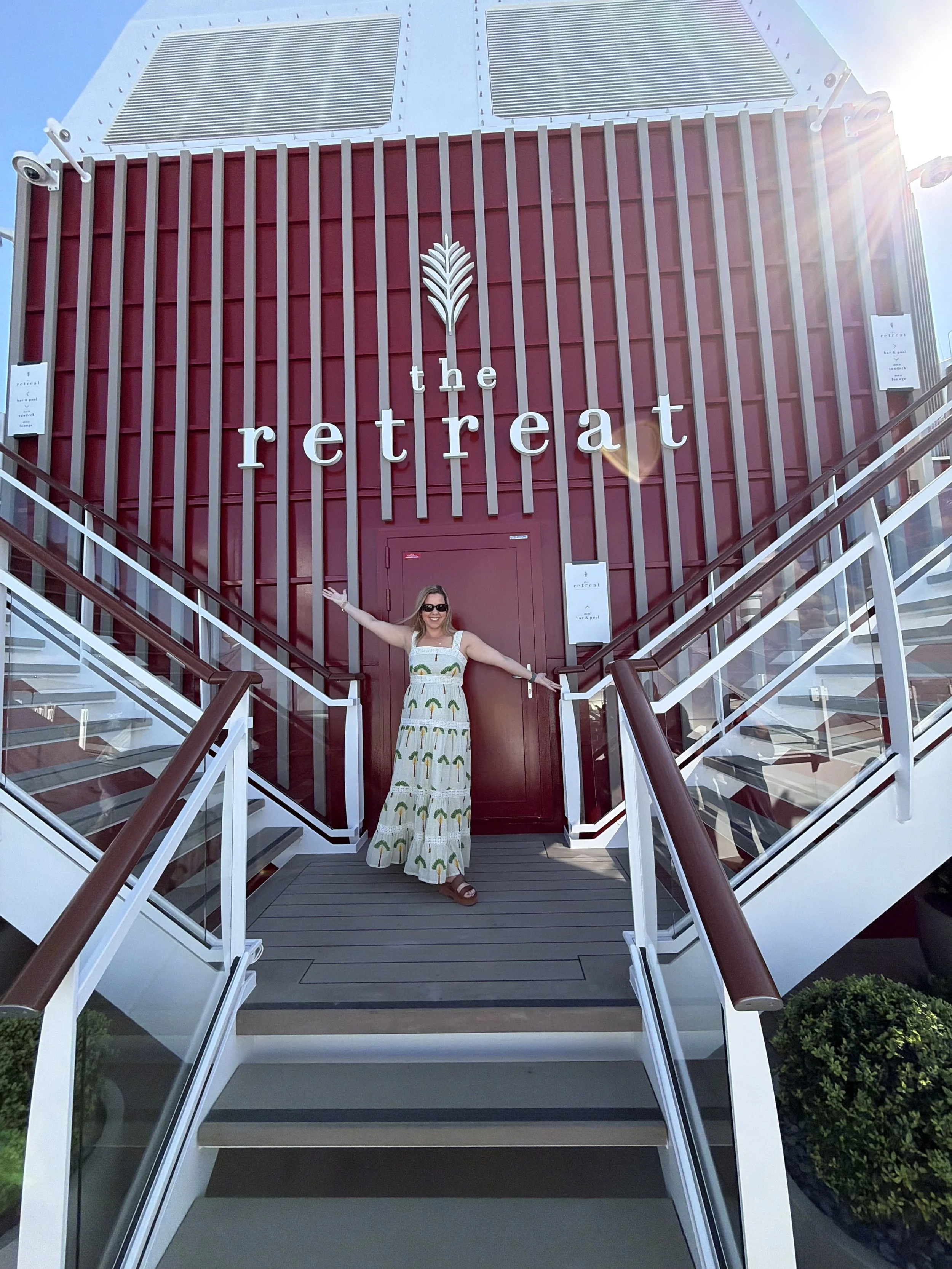 Morgan in a colorful dress with a pattern of rainbows stands on outdoor stairs with glass railings, arms outstretched, in front of a building with a red facade and large white lettering that reads 'the retreat'.