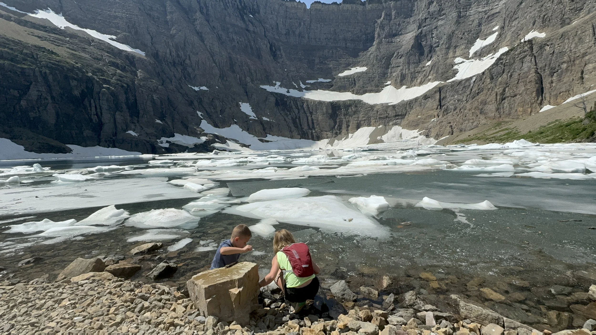 Joy and Jed, sit on rocks at the edge of a glacier-fed lake, with floating ice chunks and a towering rocky mountain in the background.