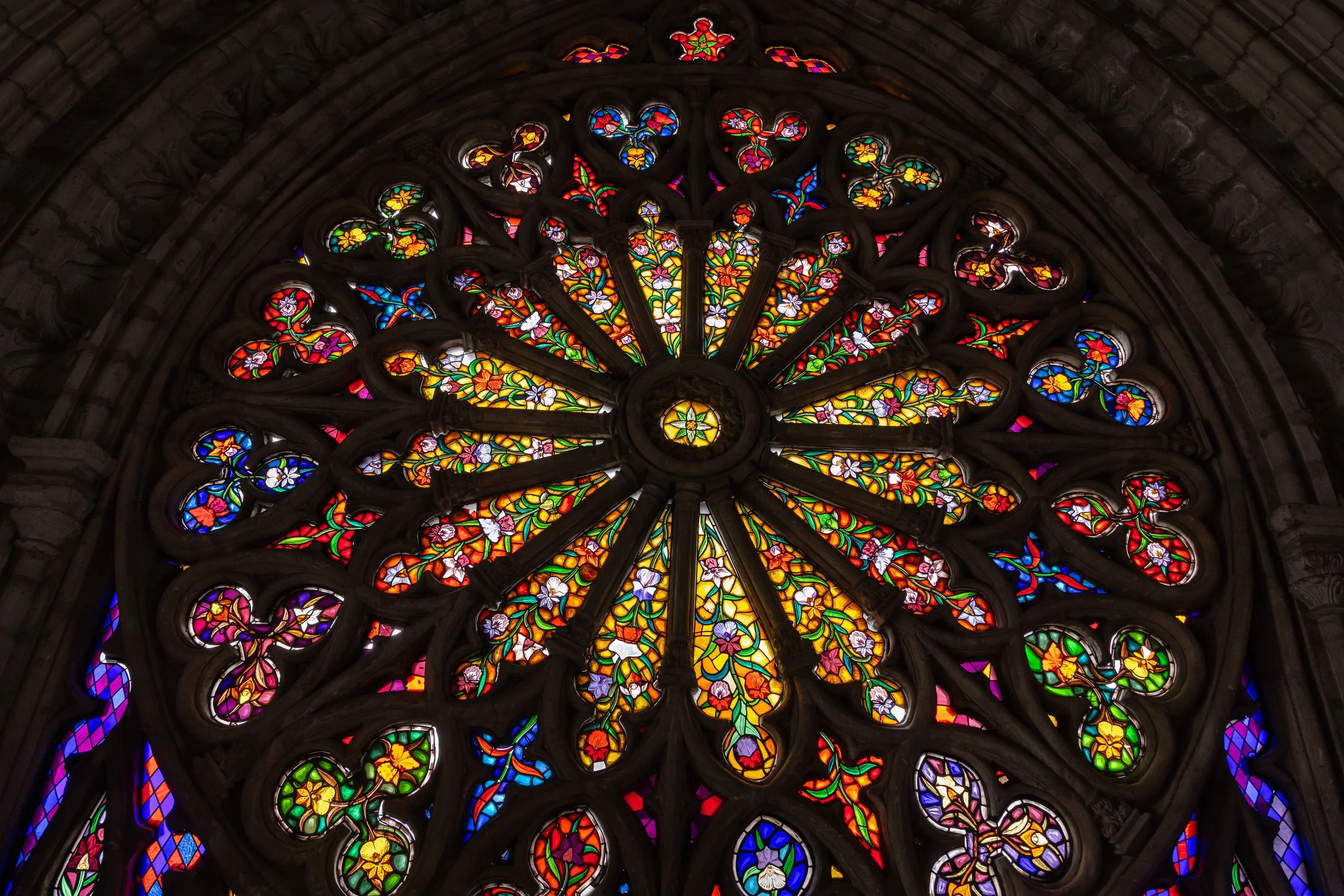 Colorful stained glass rose window with intricate floral and geometric patterns in red, yellow, blue, green, and purple hues.