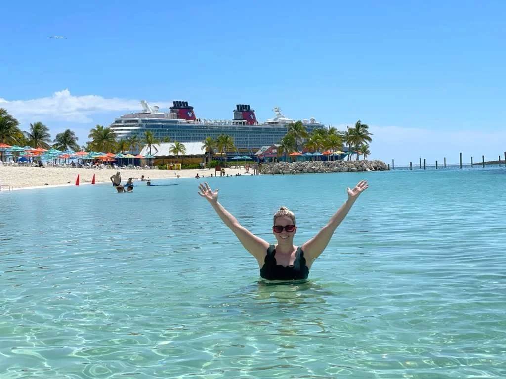 A woman in sunglasses and a black swimsuit smiling with arms raised in clear turquoise water on a sunny day, with a cruise ship, palm trees, and a beach with umbrellas in the background.