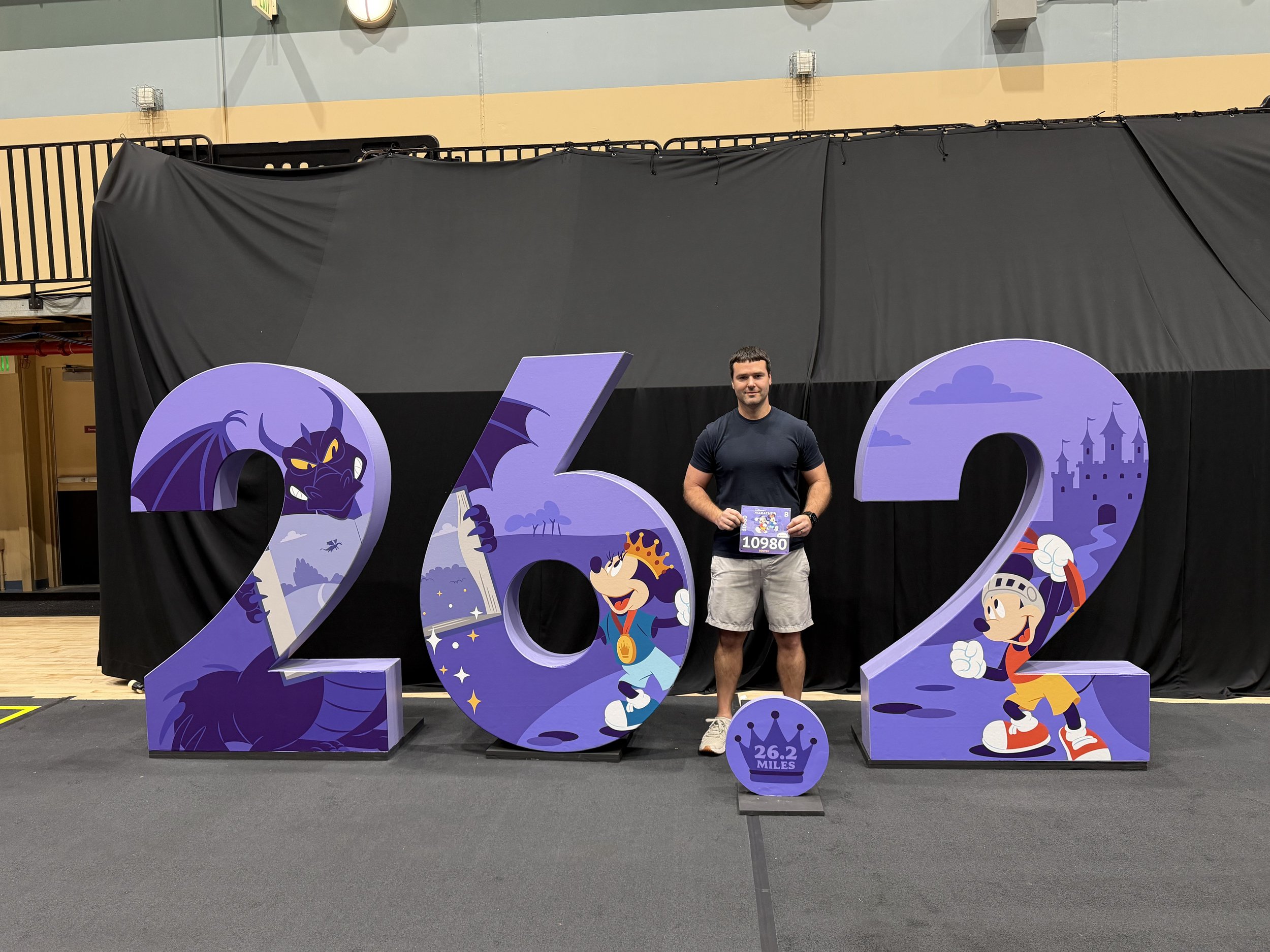 Man posing at RunDisney expo with 26.2 sign and race bib prior to running the race.