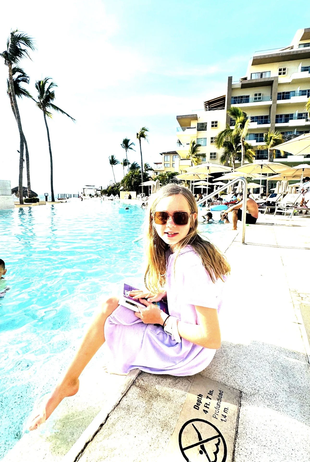Young girl, in a pool cover up, hair down, sunglasses, sitting on the side of a pool at Dreams Vallarta Bay in Puerto Vallarta, with a book in hand 
