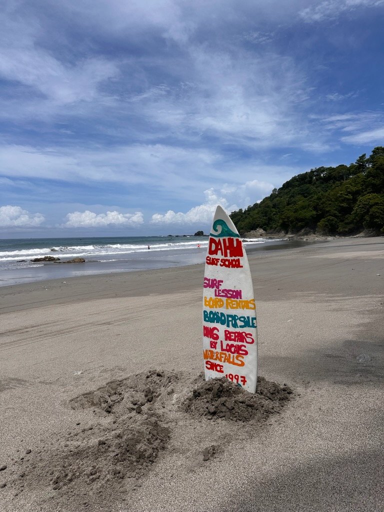 A colorful surfboard stuck in the sand on a beach with ocean waves, a green hillside, and cloudy sky in the background.