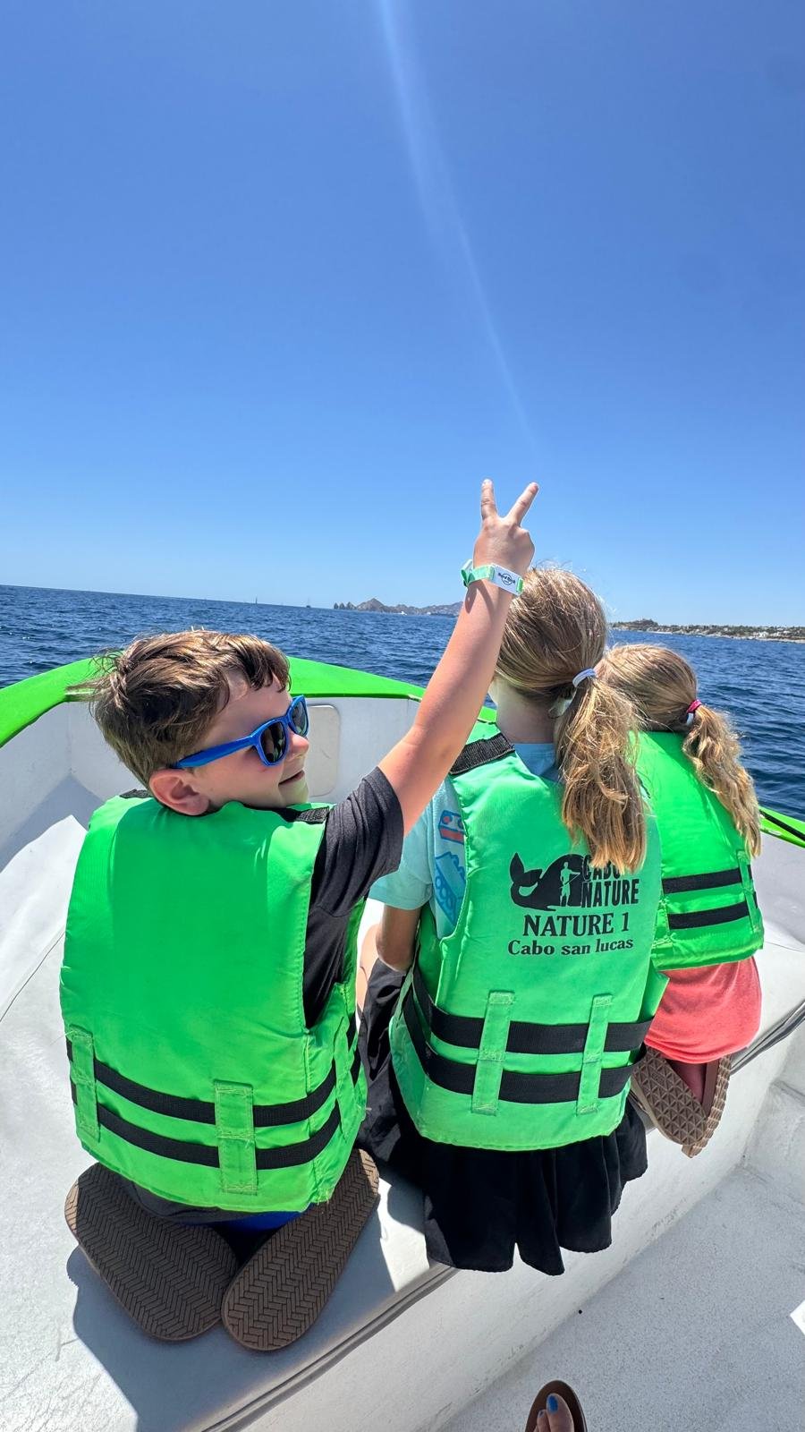 Sanders Kids wearing green life jackets sitting on a boat, with the boy in sunglasses raising his hand in a peace sign.