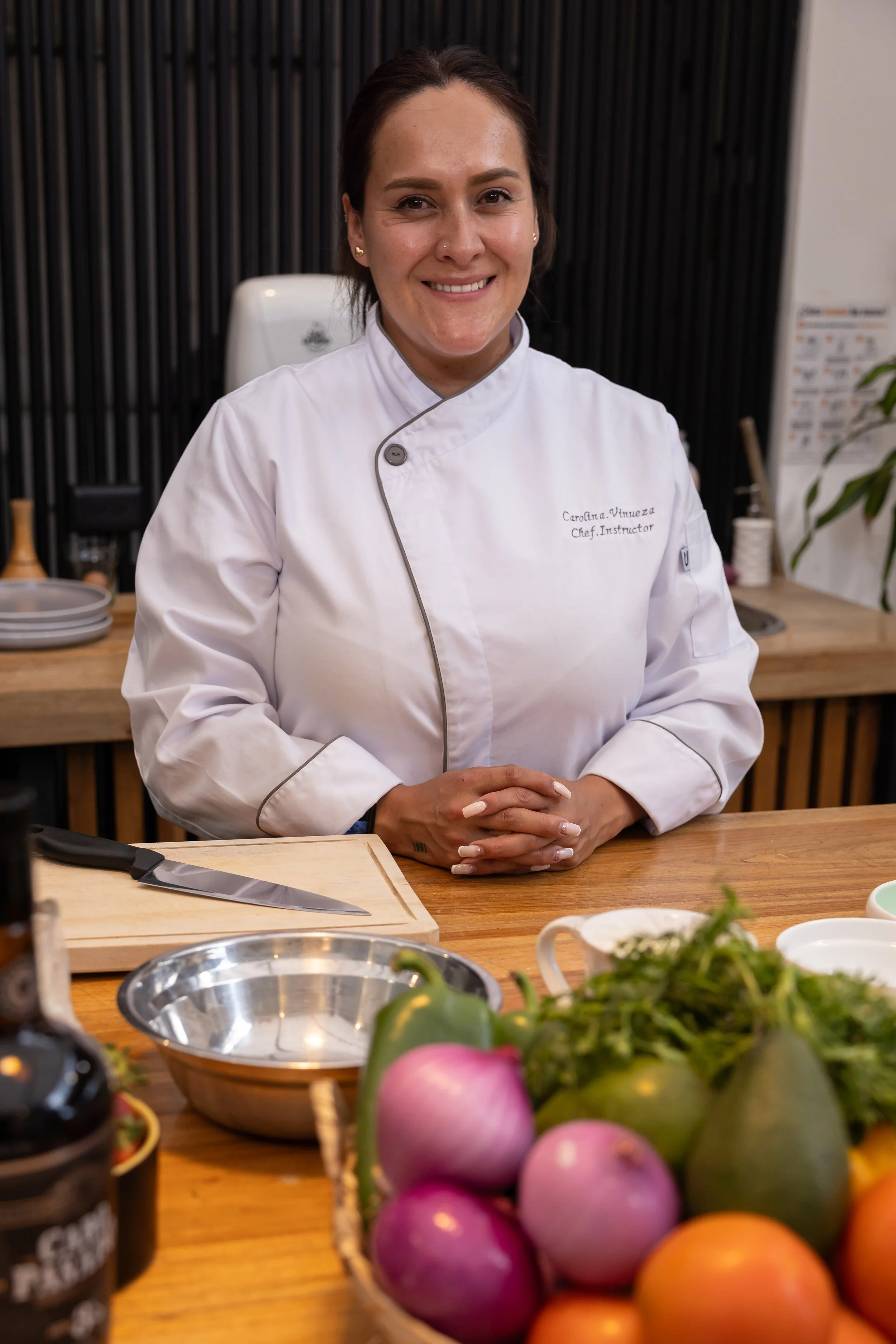A female chef smiling in a professional kitchen, wearing a white chef's coat with her name and title embroidered on it, seated at a wooden counter with various cooking ingredients and utensils around her.