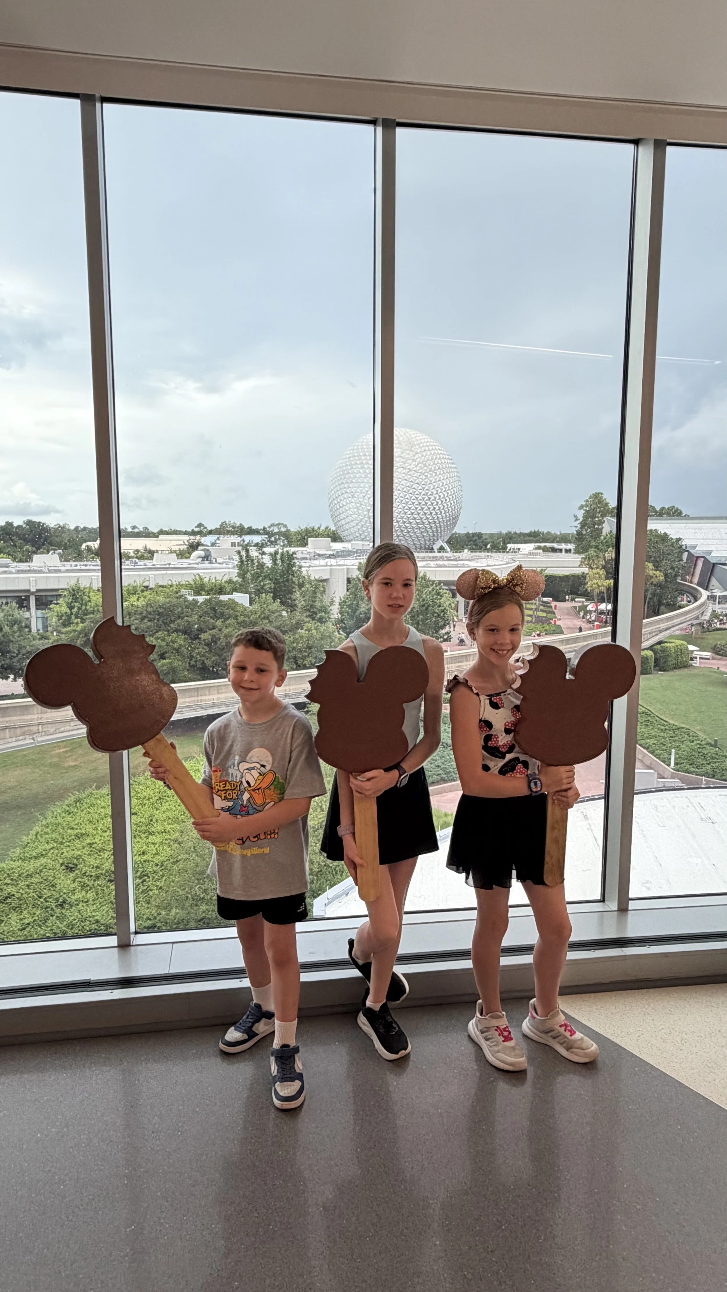 Sanders kids standing in front of a large window with a view of Geodesic Sphere and Disney World background, holding Disney-themed Mickey Mouse shaped signs.