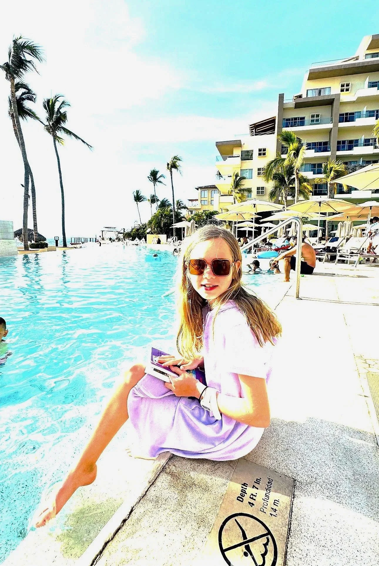 Young girl, in a pool cover up, hair down, sunglasses, sitting on the side of a pool at Dreams Vallarta Bay in Puerto Vallarta, with a book in hand 