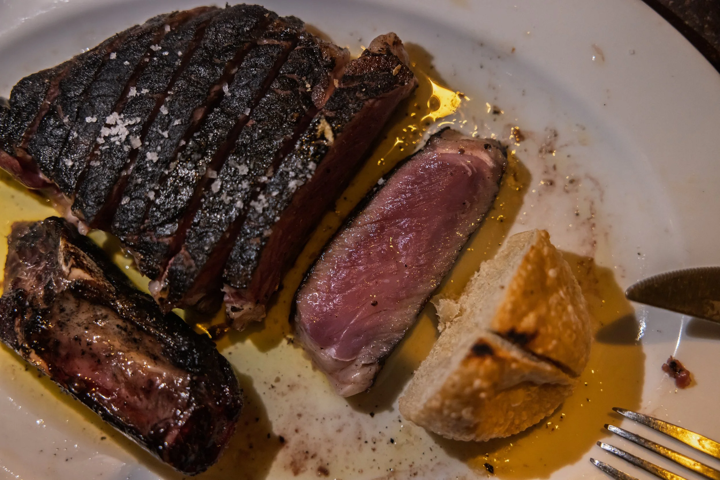 A plate with slices of cooked steak, with a charred crust, some salt on top, and surrounded by pink interior. There is a piece of bread and some fried food, with a fork on the side. Olive oil drips from the meat, and the plate has some oil and season