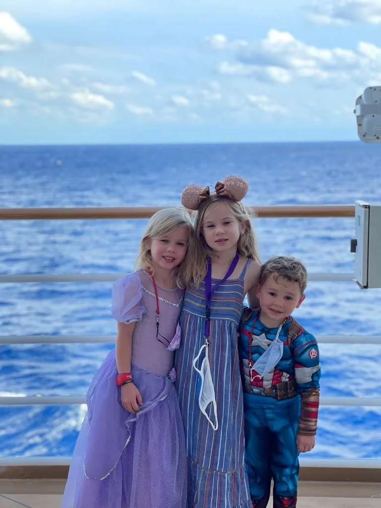 Three children standing on a ship deck with an ocean background, smiling at the camera. They are dressed casually and have face masks hanging from their necks.