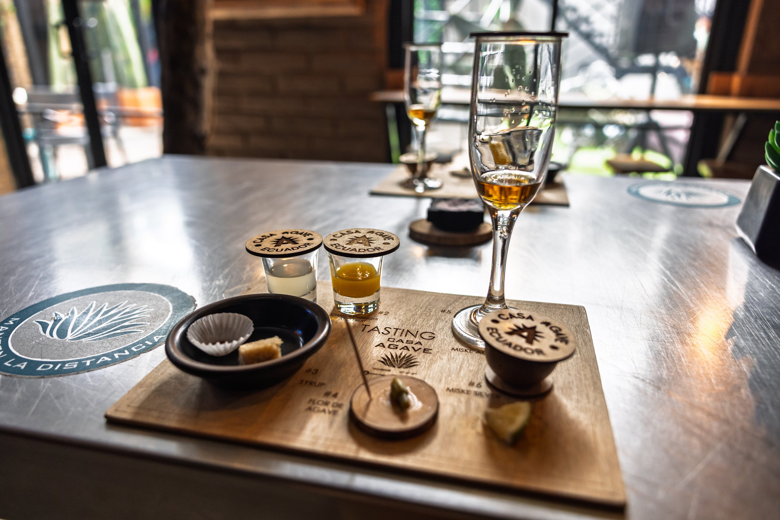 Close-up of a table with small samples of agave syrup on wooden coasters, a glass of water with ice and a lemon wedge, and a tasting sheet in the background at Casa Aguamiel distillery.