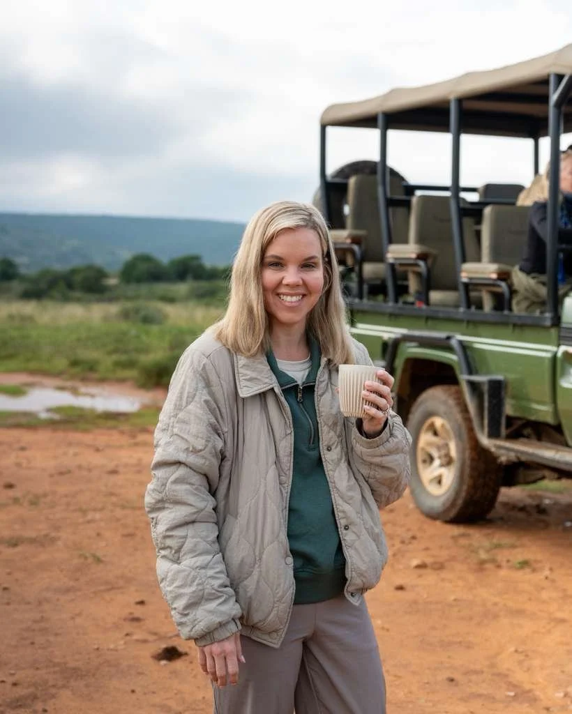 Morgan smiling and holding a white mug outdoors, standing on a dirt area with a safari vehicle and green landscape in the background.
