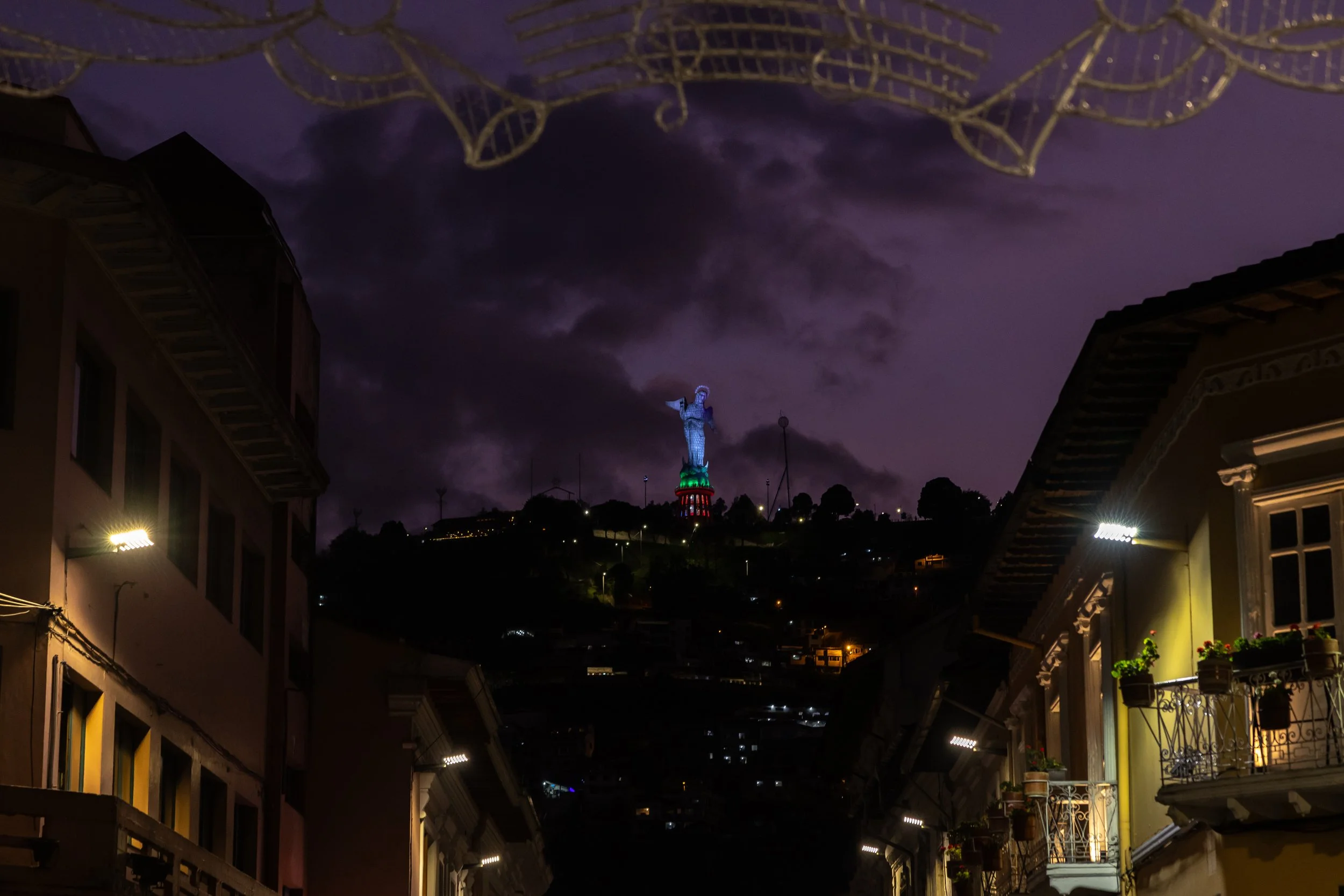 Night scene of a city with buildings, a hill with a lit-up statue, and a dark purple sky with clouds and visible electrical wires.