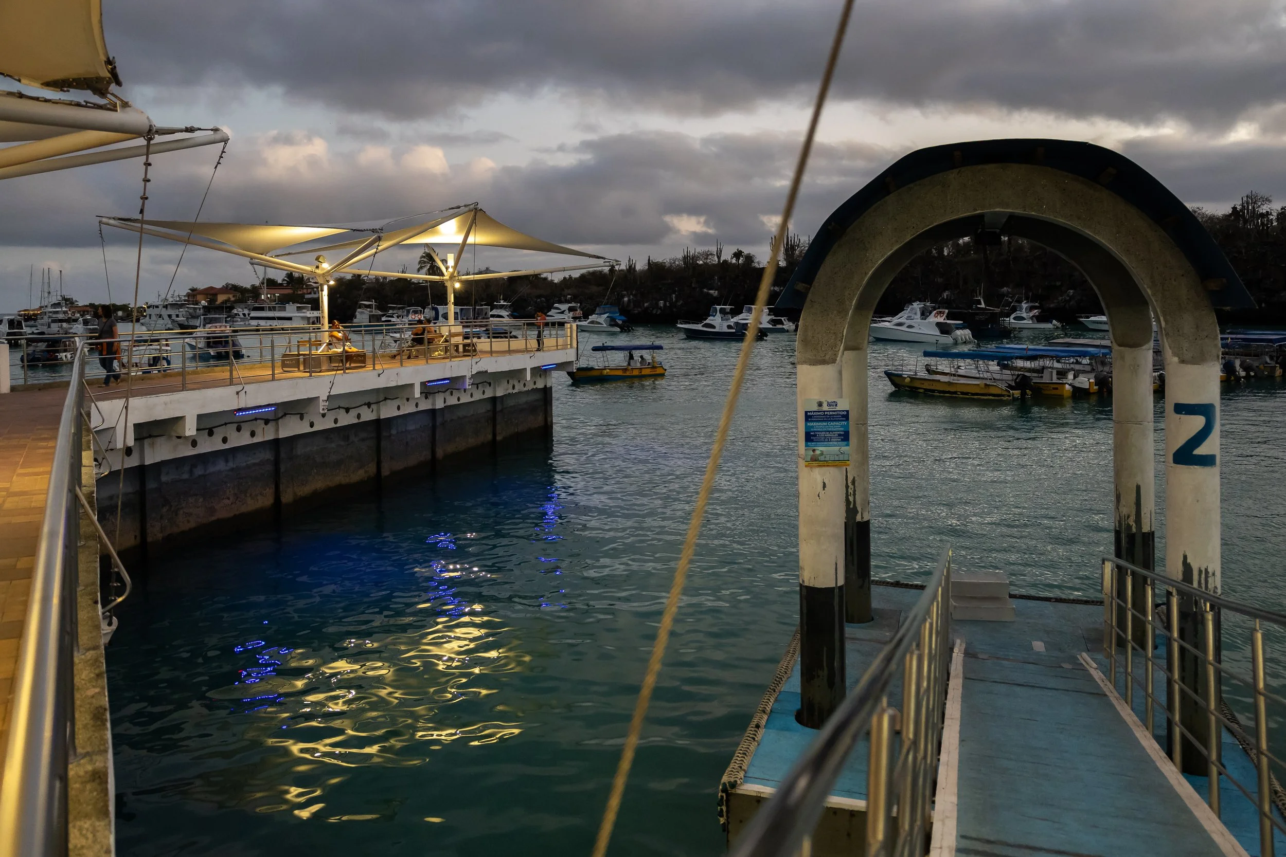 A marina at dusk with boats docked in the water, a pier with people, and a cloudy sky overhead.