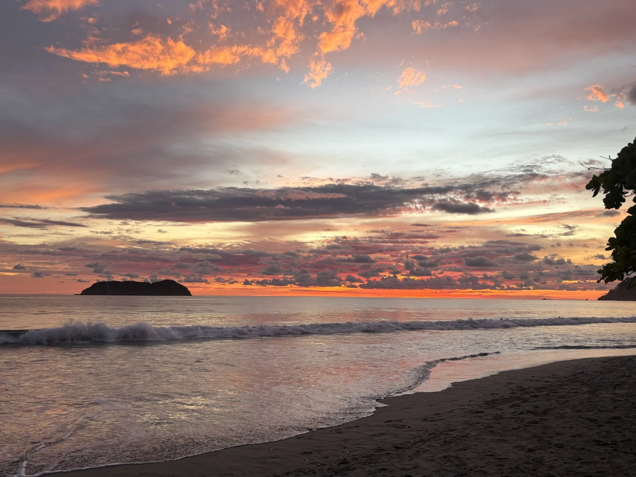 Sunset over the ocean with a partly cloudy sky and a small island in the distance. The beach is in the foreground with waves gently washing ashore.
