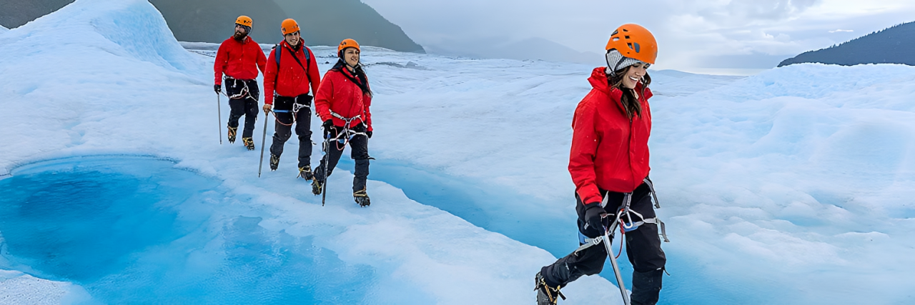Group of four climbers wearing red jackets and orange helmets walking on icy terrain in a glacier landscape.