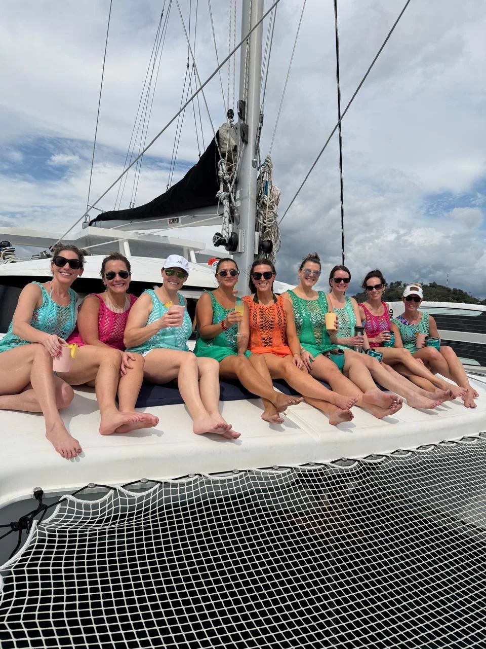 Group of nine women sitting on the deck of a boat, enjoying drinks, under partly cloudy skies.