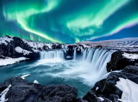 Northern lights over a waterfall in a snowy landscape with dark rocks.