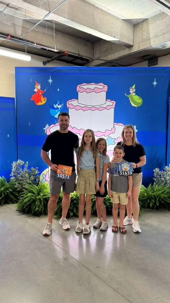 A man and woman standing with three young girls in front of a colorful backdrop with a tiered cake and fairy tale characters, suggesting a celebration or event.