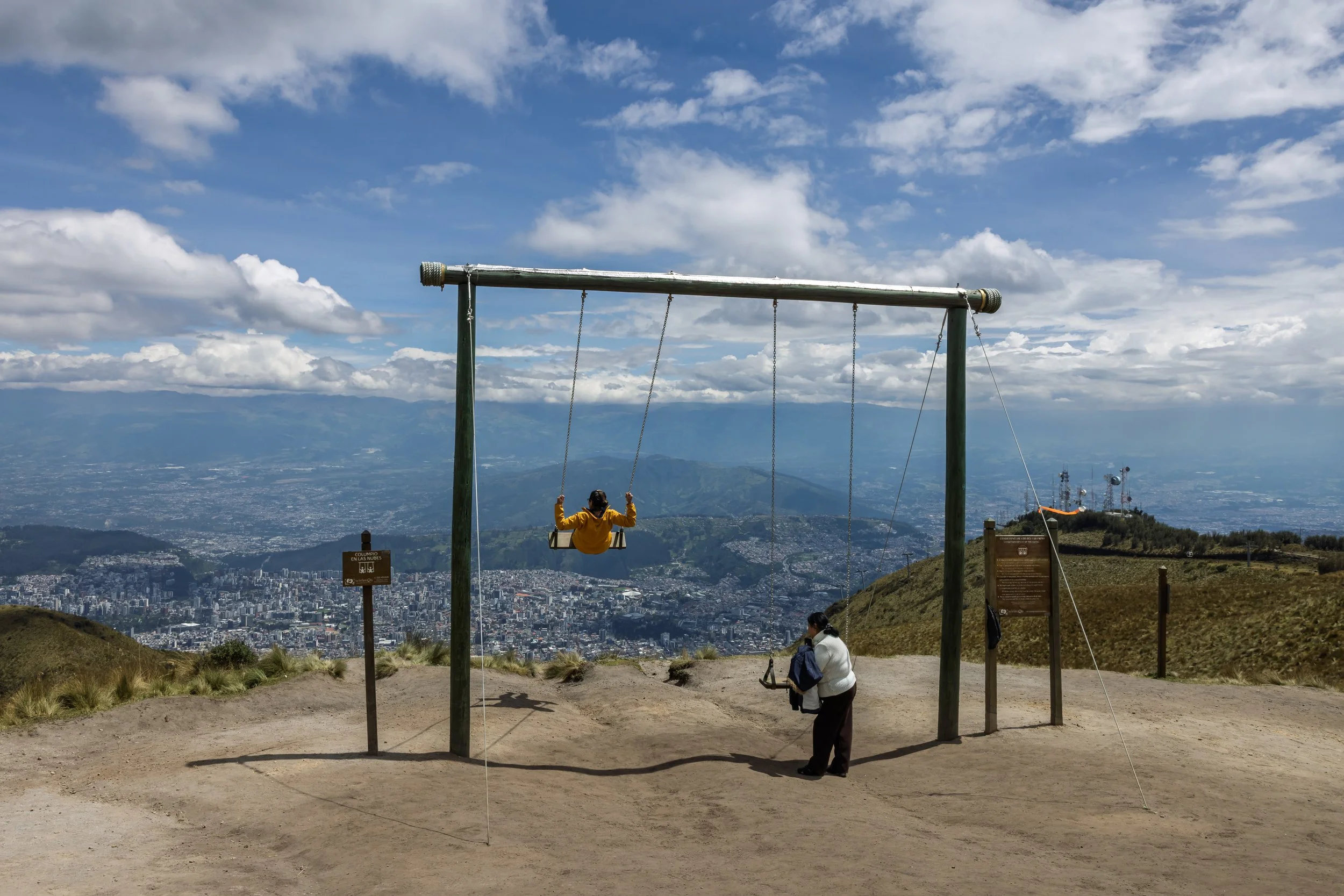A person in a yellow jacket is sitting on a swing hanging from a large wooden frame at a mountain viewpoint, with a panoramic view of a city and surrounding mountains under a partly cloudy sky.