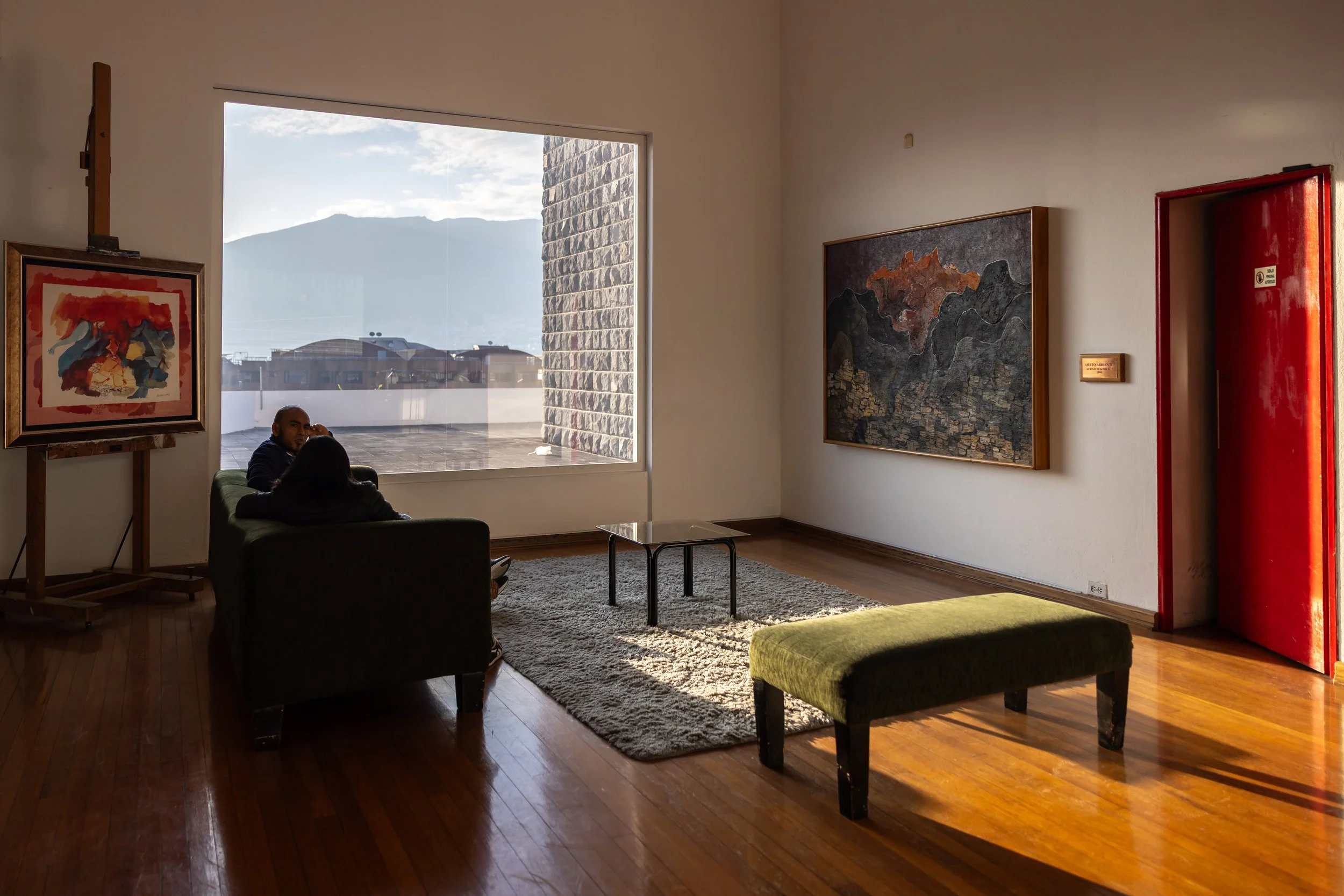 An art gallery interior with paintings on white walls in front of a large window with a mountain view. Furniture includes a green chair, a small table, and a green ottoman. A person is sitting on the chair, partially visible. The floor is wooden, and