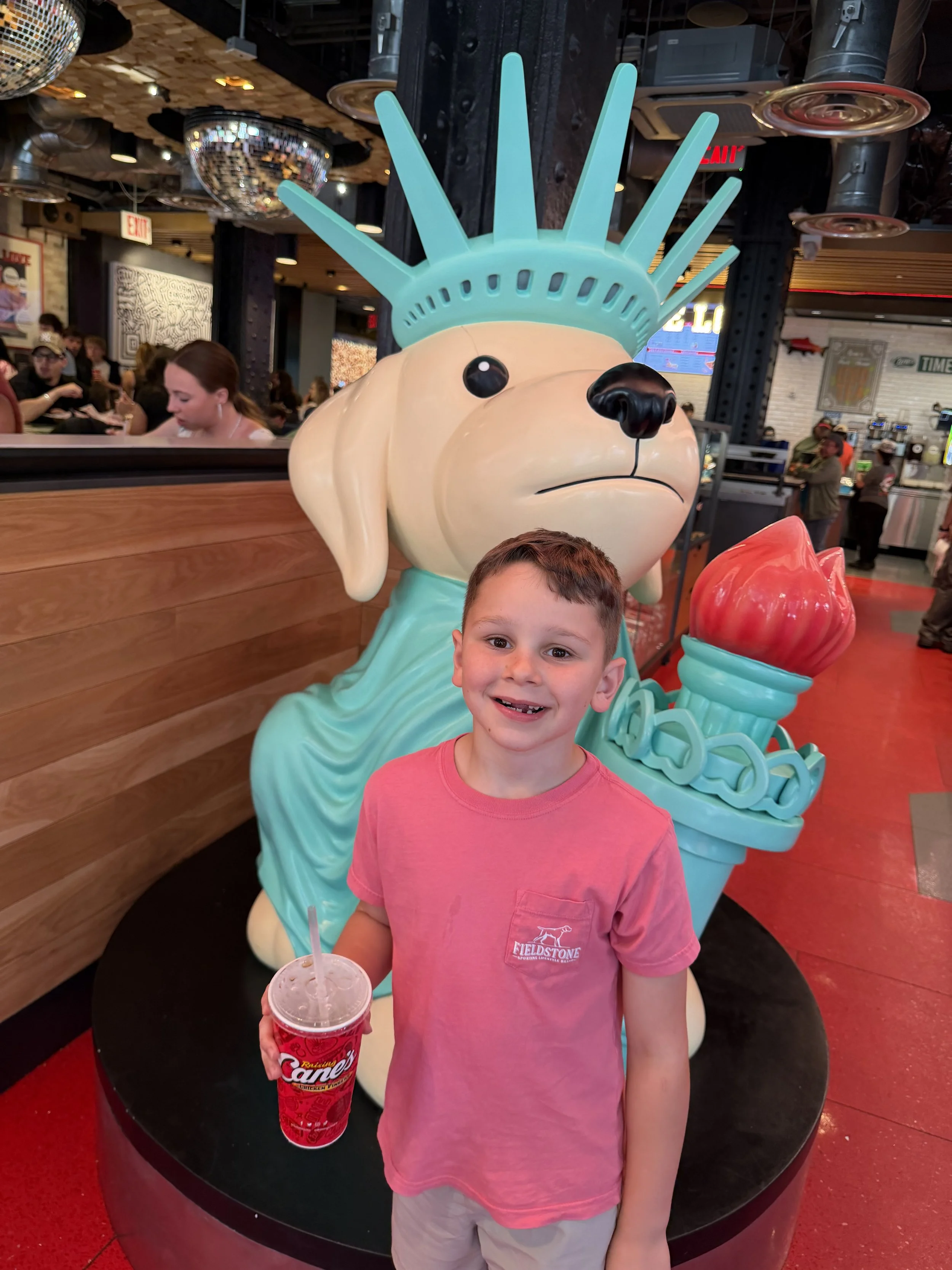 Jed holding a drink stands in front of a large statue of the Statue of Liberty's head inside a restaurant or cafe.