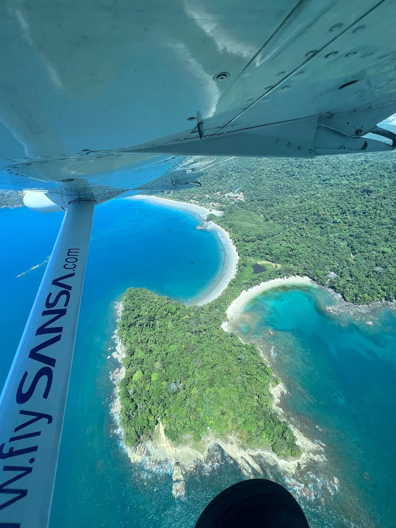 An aerial view from an aircraft showing the coastline with a beach, greenery, and blue waters with two small lakes or lagoons near the shore.