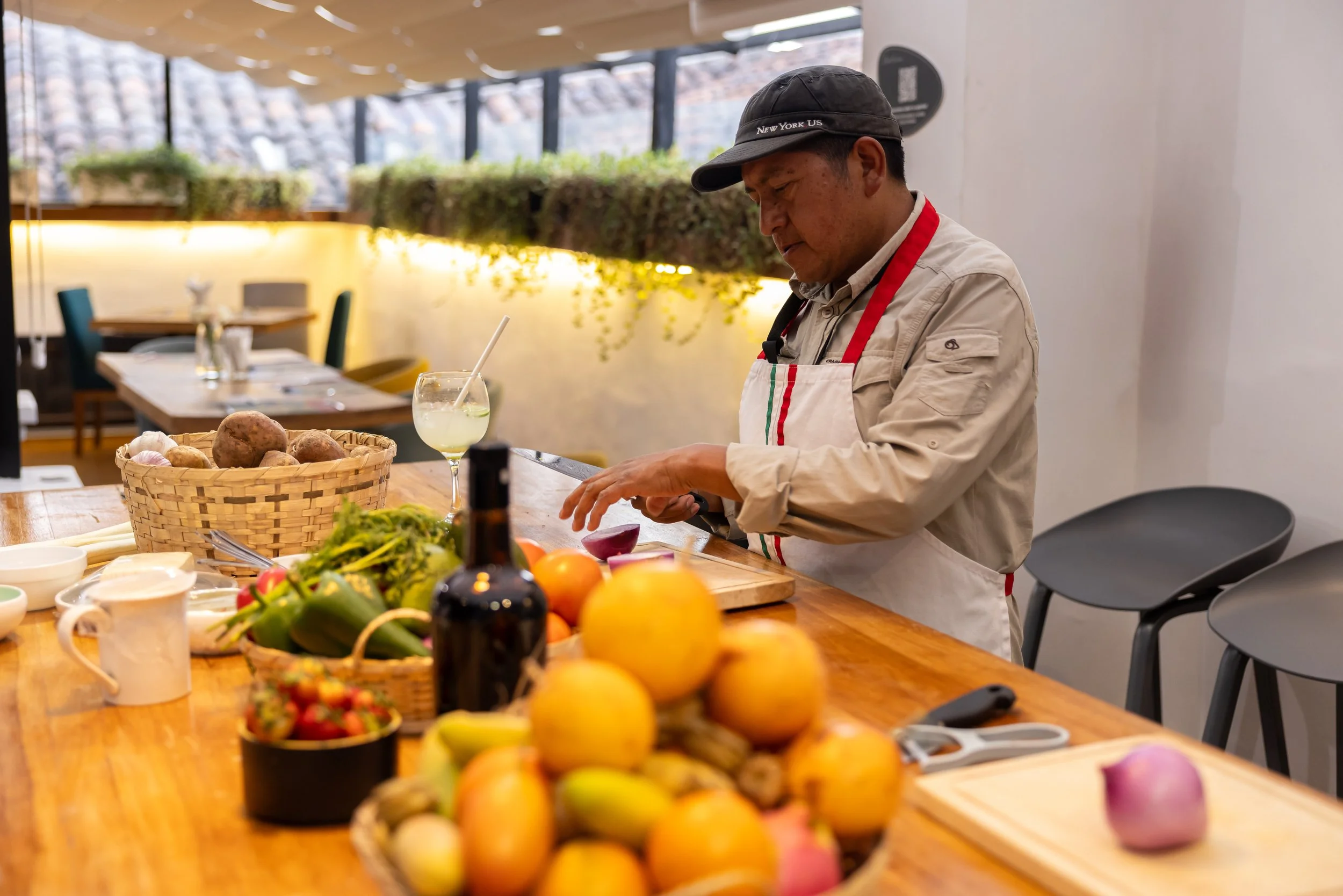 A man wearing a black cap and beige chef's coat stands at a wooden table filled with fresh vegetables and fruits, including potatoes, tomatoes, lemons, and strawberries, in a bright, modern restaurant with large windows and green plants in the backgr