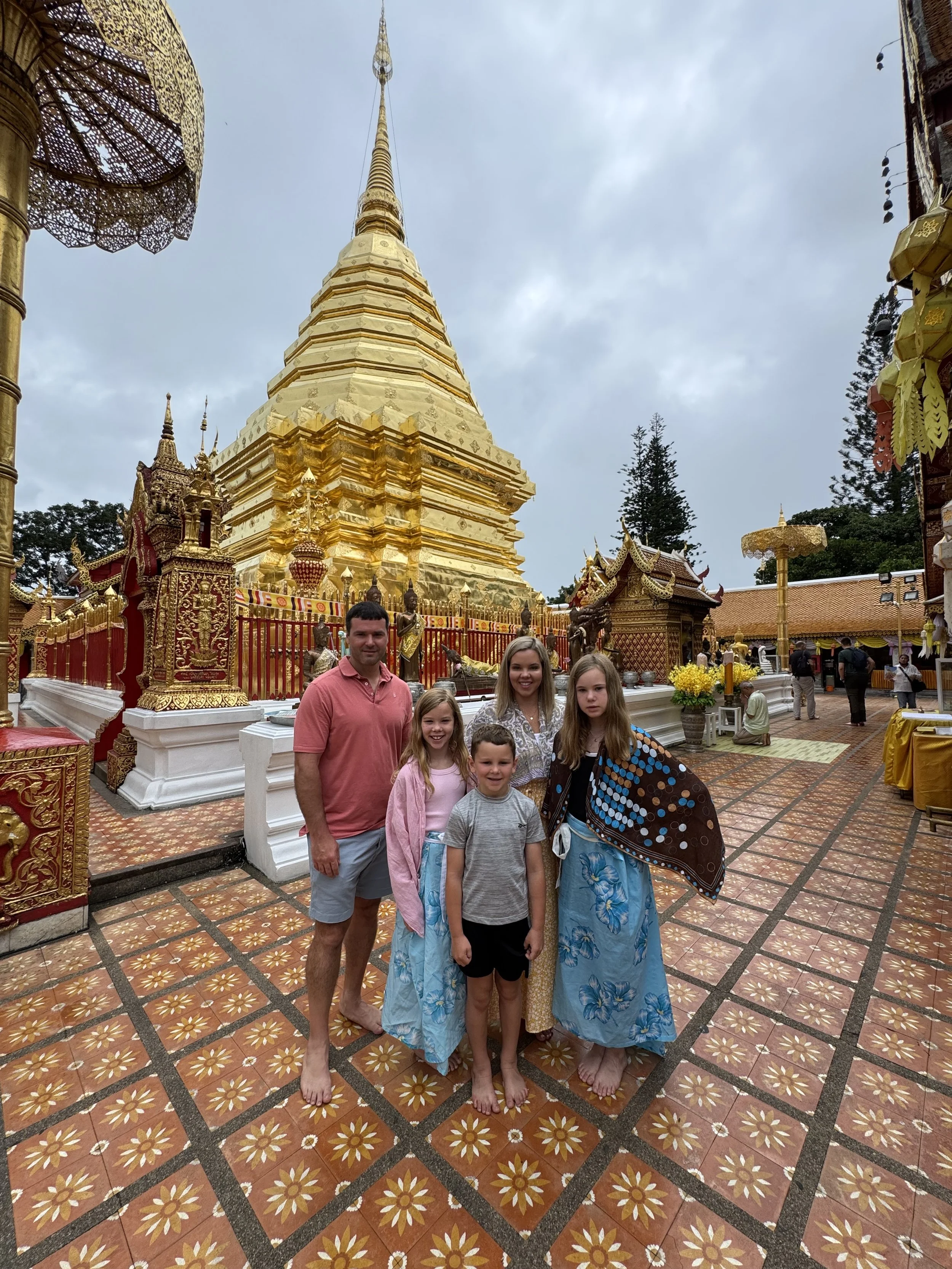 Sanders Family, standing barefoot in front of a golden religious structure at a temple. The background shows ornate decorations, statues, and small temple buildings under a cloudy sky.