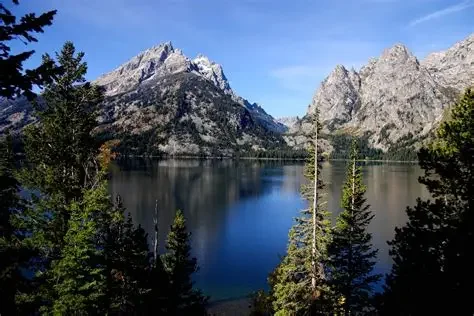 A clear lake surrounded by pine trees with rugged mountains in the background under a blue sky.