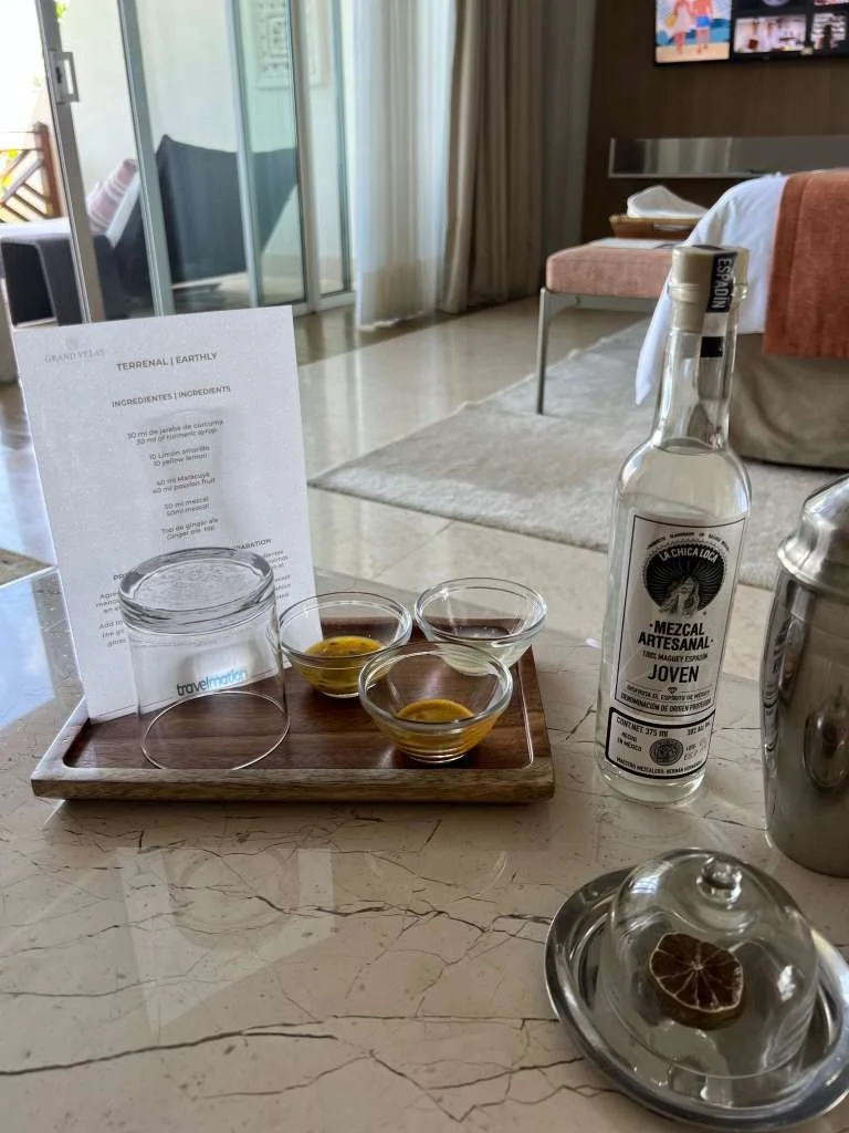 Hotel room with glass sliding door, curtains, with Three small bowls of tequila and lime slices, a mezcal bottle, and a note on a marble countertop.