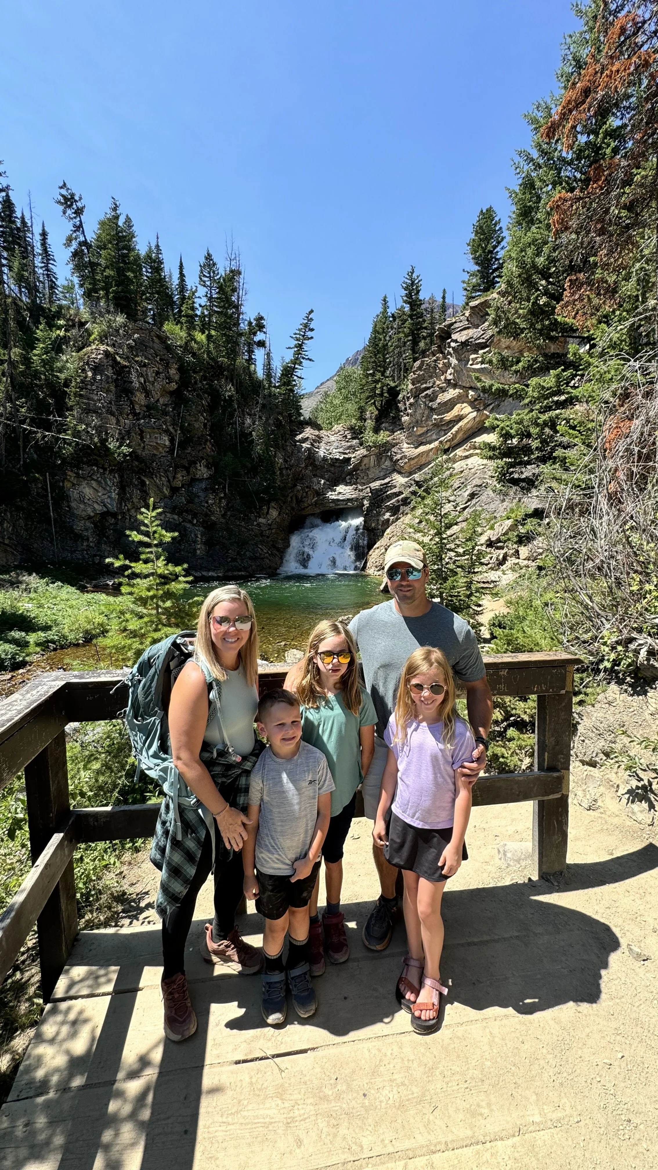 Sanders Family standing on a wooden bridge in front of a waterfall in a forested mountain setting on a sunny day