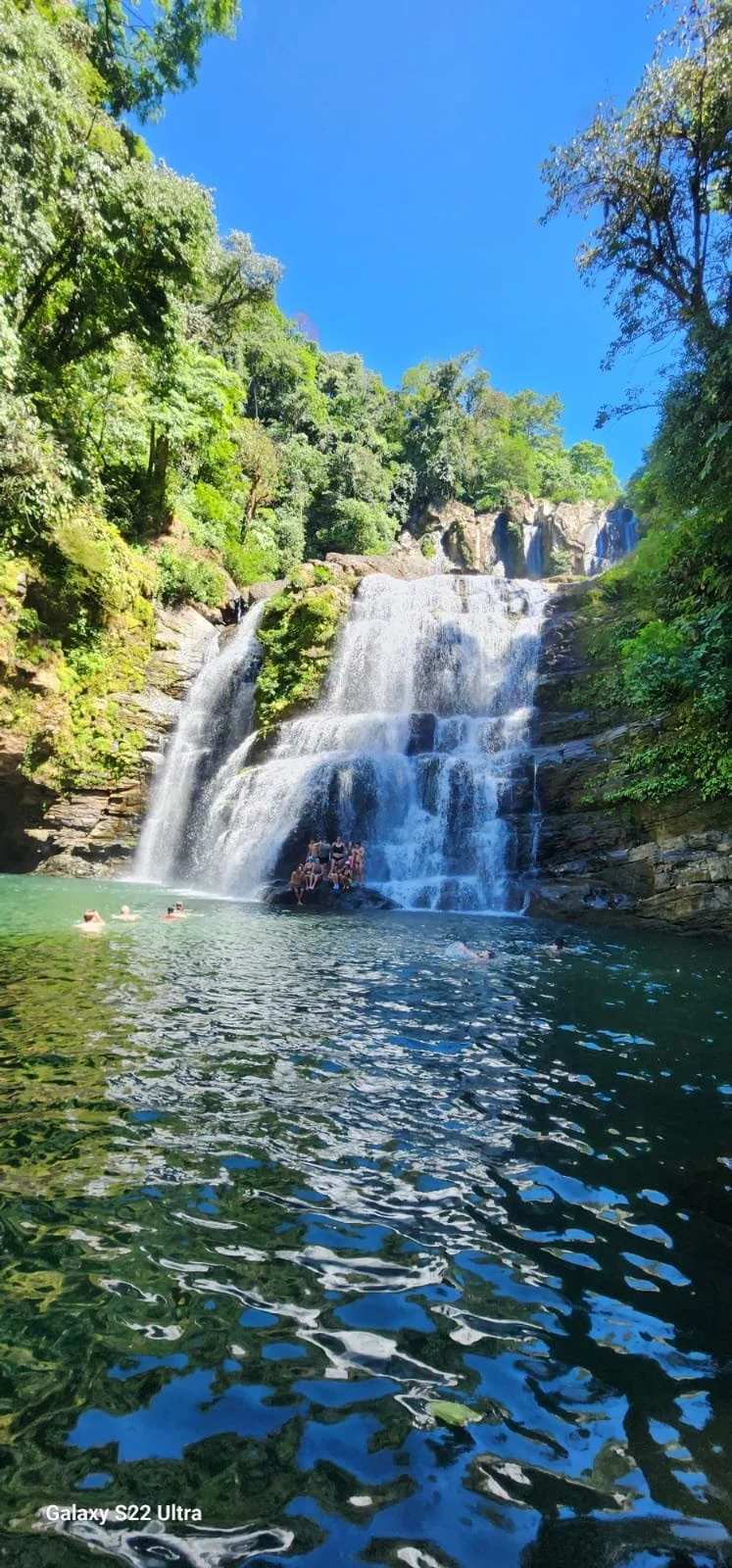 Waterfall cascading into a pool surrounded by lush green trees under a clear blue sky with a few people swimming and relaxing in the water.