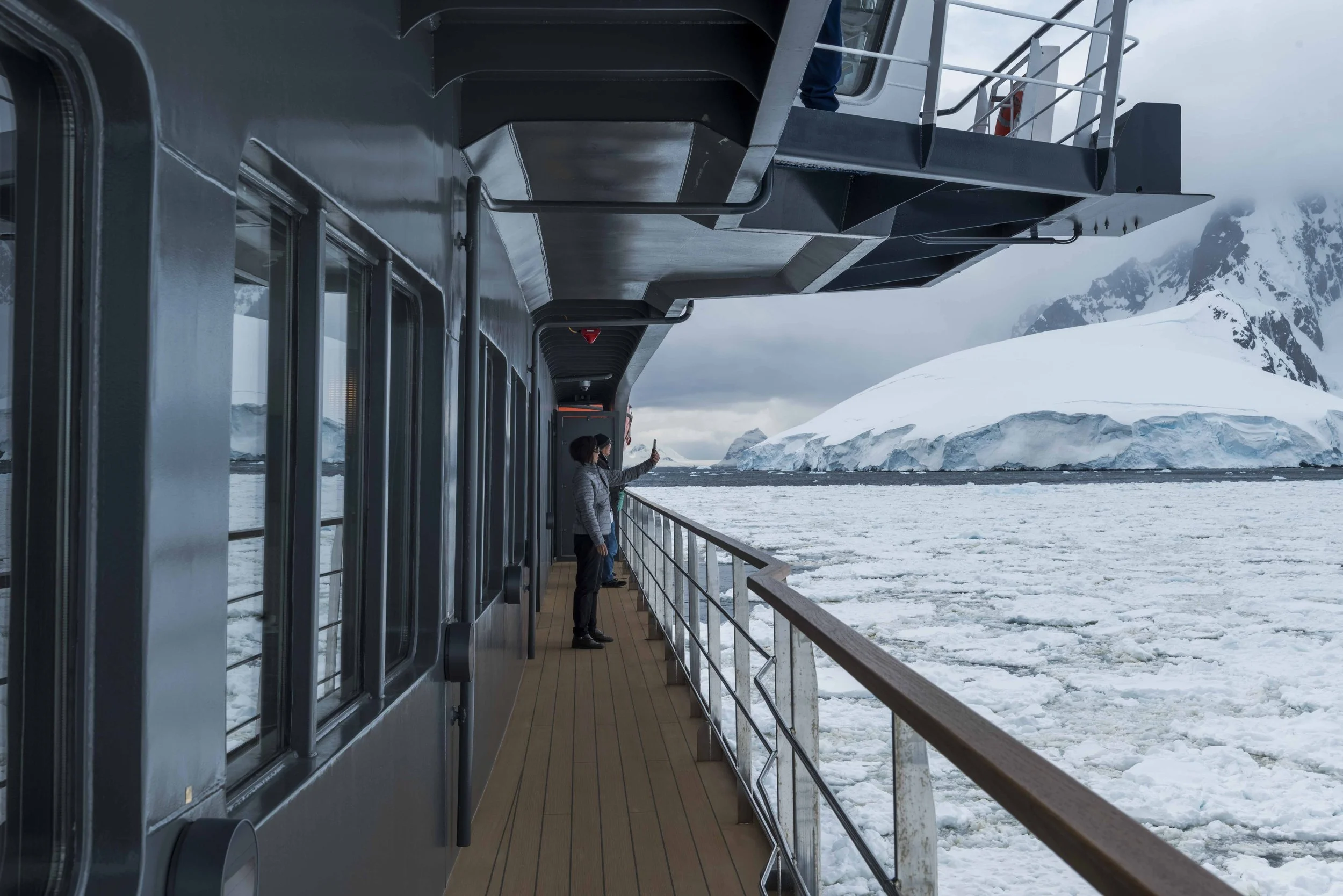 People standing on the deck of a ship in icy waters with snow-covered mountains in the background.
