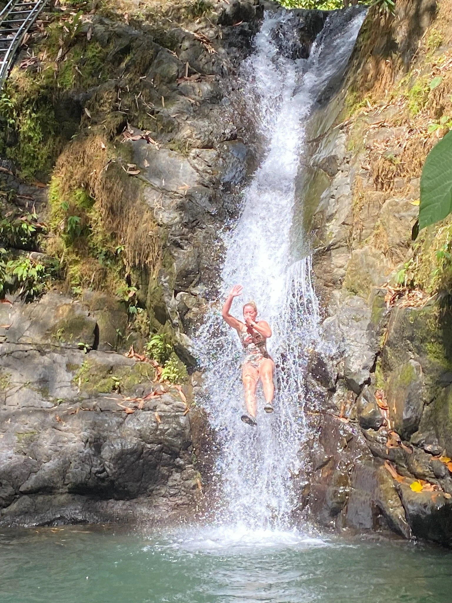 A person in a swimsuit standing in a small waterfall or stream in a natural outdoor setting with rocks and greenery.