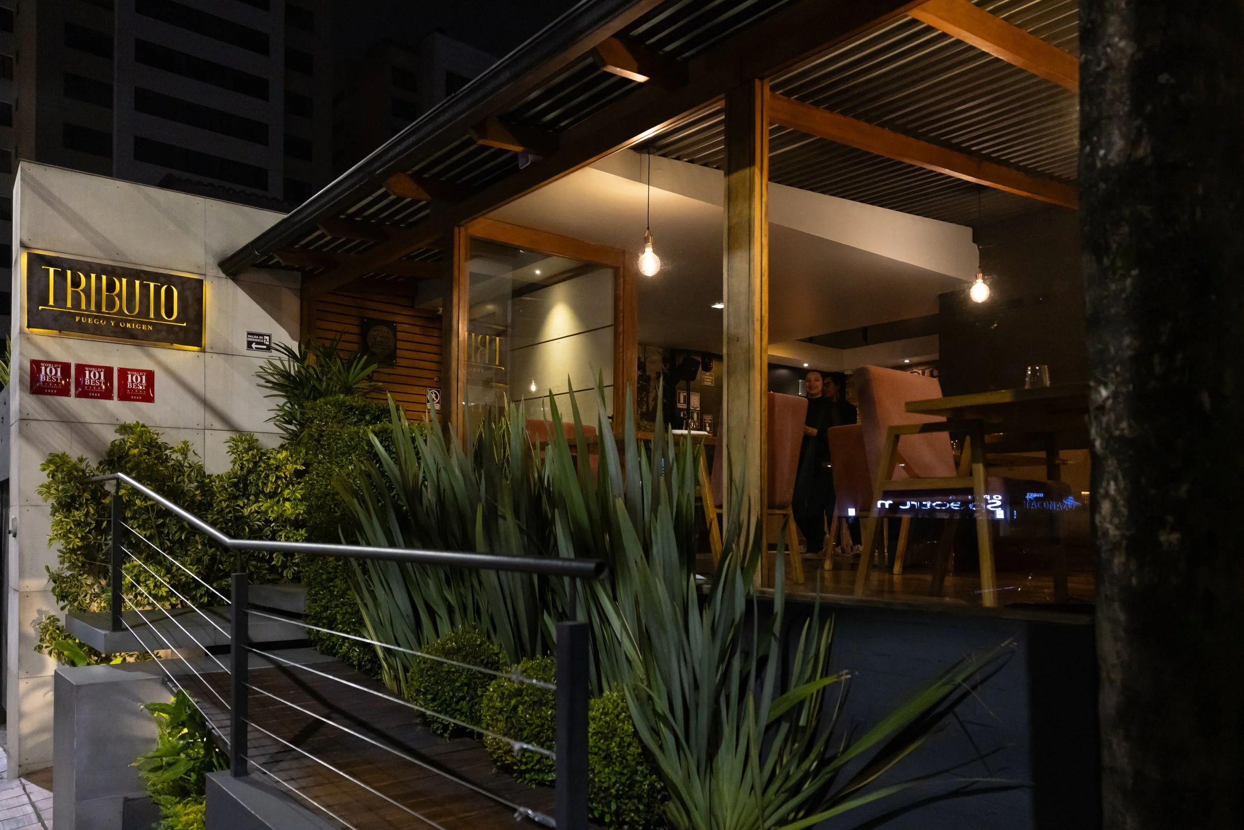 The image shows an outdoor restaurant or cafe area at night with warm lighting. There are plants in the foreground, a wooden deck, and a structure with hanging light bulbs. People can be seen inside, and there are signs on the wall, including a yello