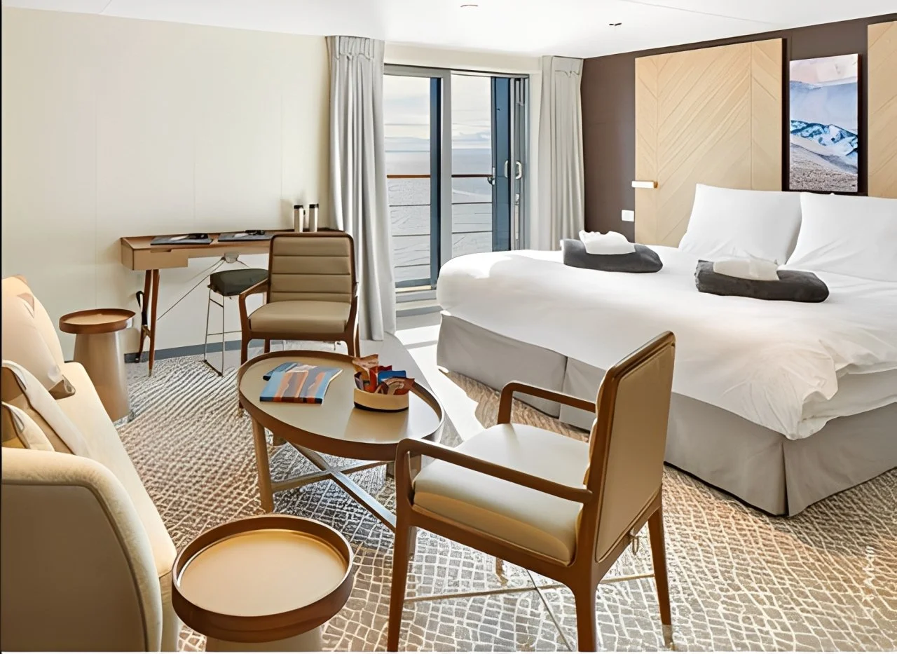 A hotel room with a king-sized bed, a balcony with ocean view, a small table with chairs, a desk, and modern decor.