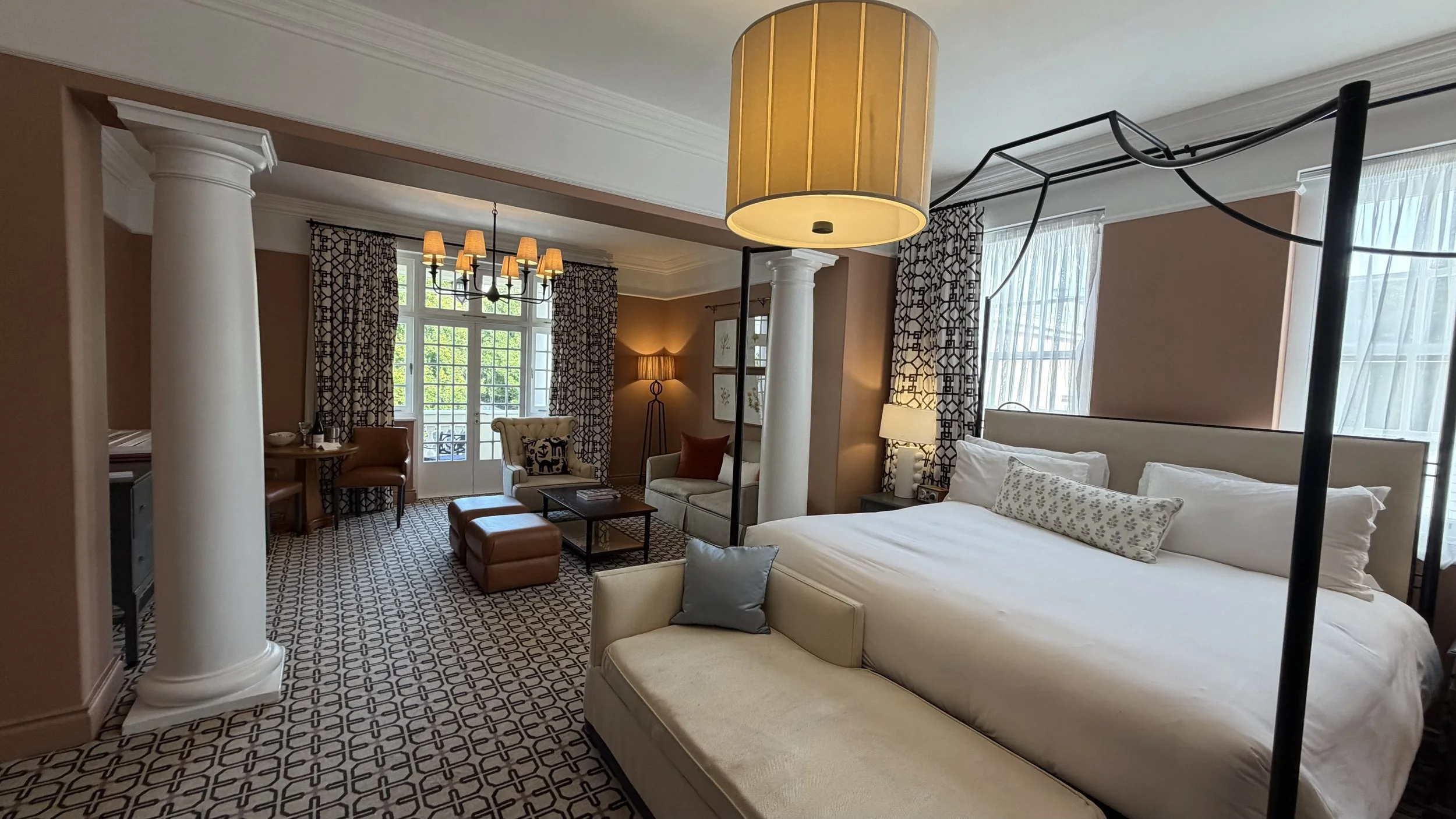 A hotel room with a bed, sitting area, and large windows, decorated with patterned curtains and warm lighting.