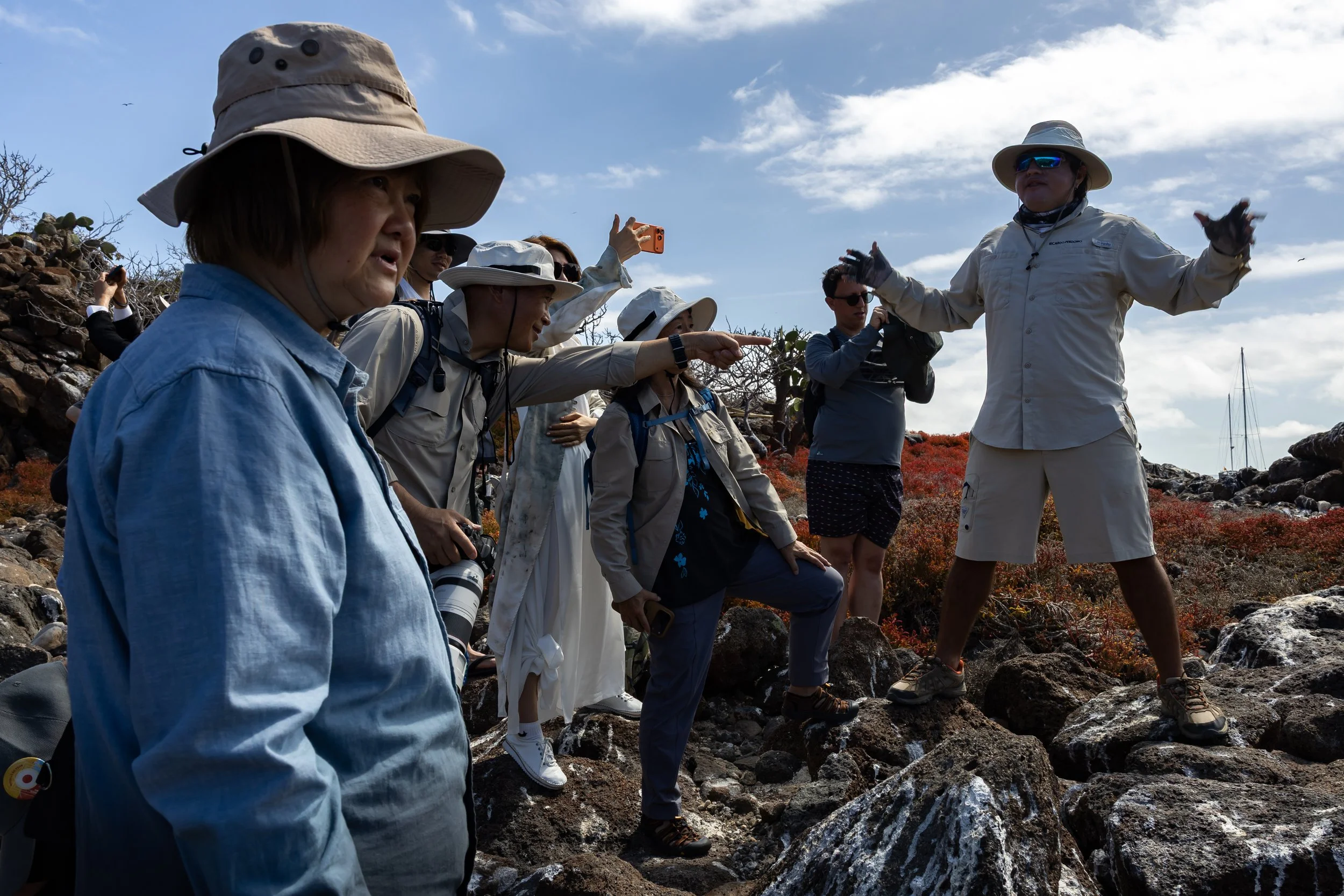 Group of people wearing hiking gear and sun hats, standing on rocky terrain outdoors, with some taking photos and others engaged in conversation under a partly cloudy sky.