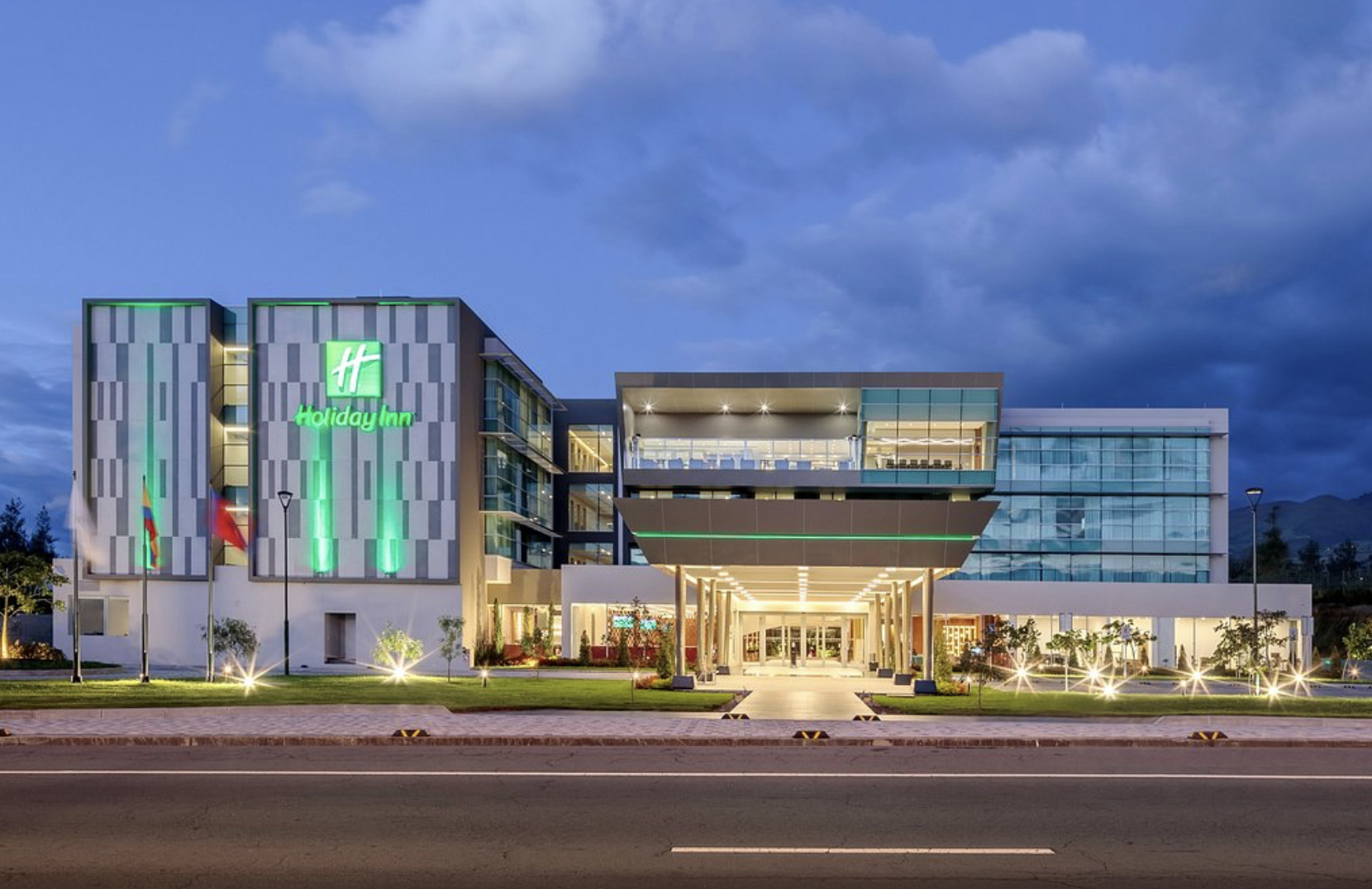 Exterior of a Holiday Inn hotel at dusk, with illuminated signage, modern glass facade, landscaping, and street lights.