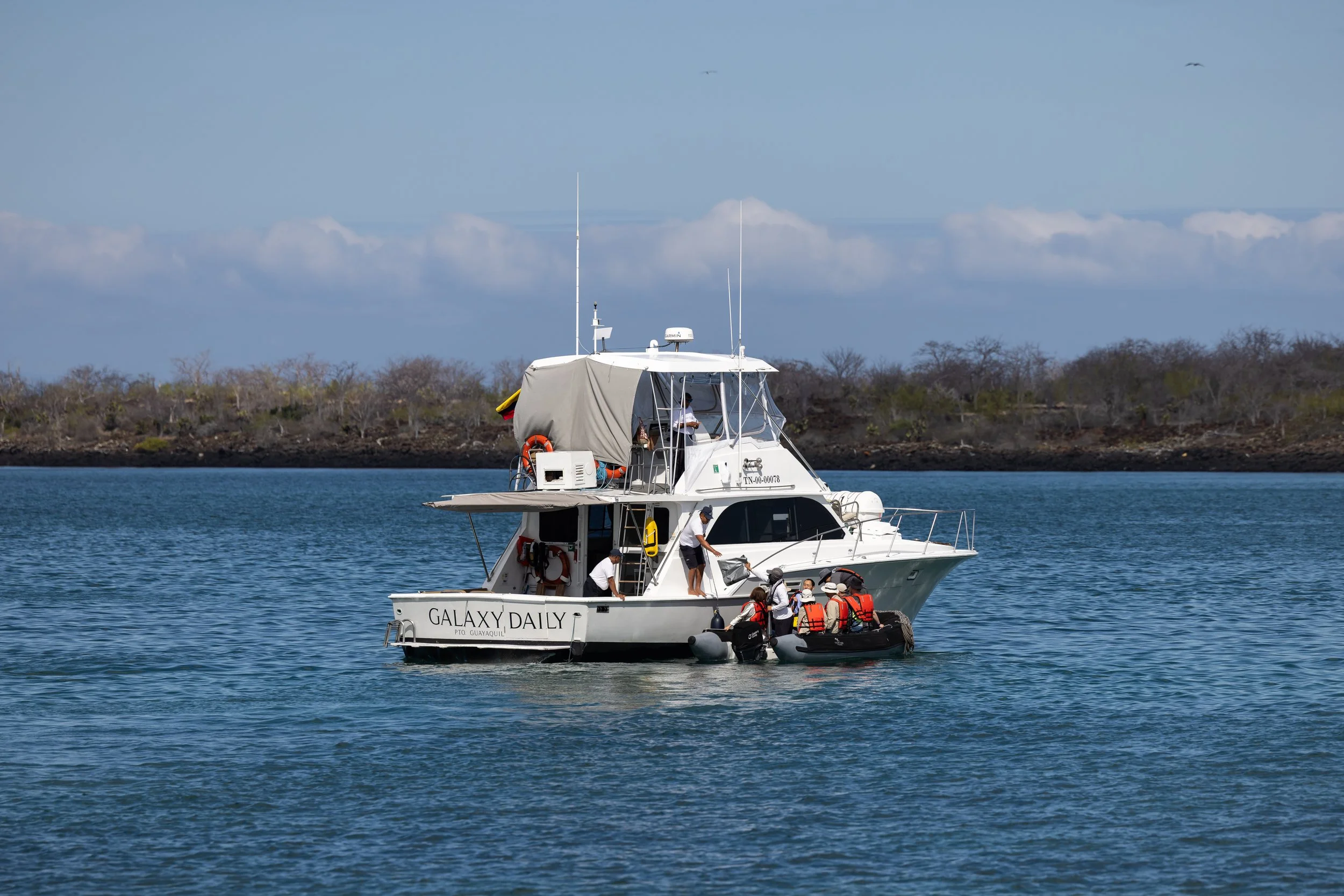 A white yacht named GALAXY DAILY with people on board and life jackets, anchored on calm water near a shoreline with trees, under a partly cloudy sky with birds flying overhead.