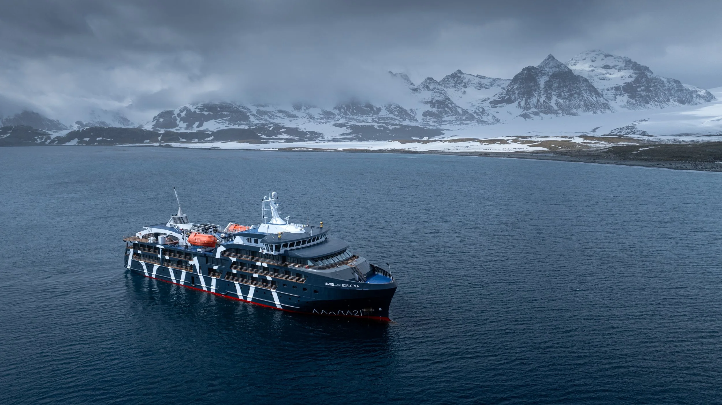 A large expedition cruise ship named Magellan Explorer sailing in icy waters with snow-covered mountains in the background under a cloudy sky.