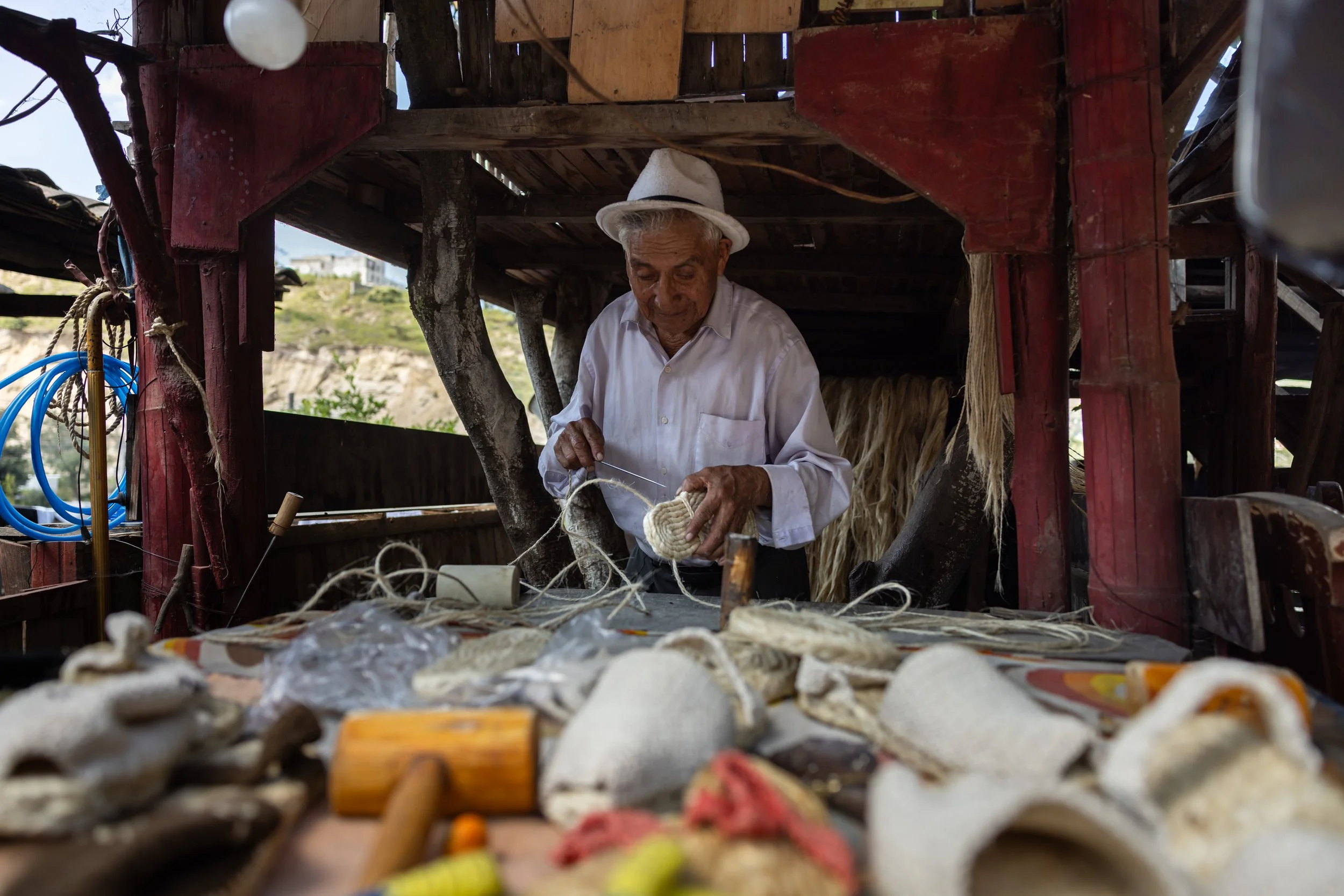 An elderly man in a white shirt and white hat tying a rope inside a rustic wooden structure with a landscape in the background.
