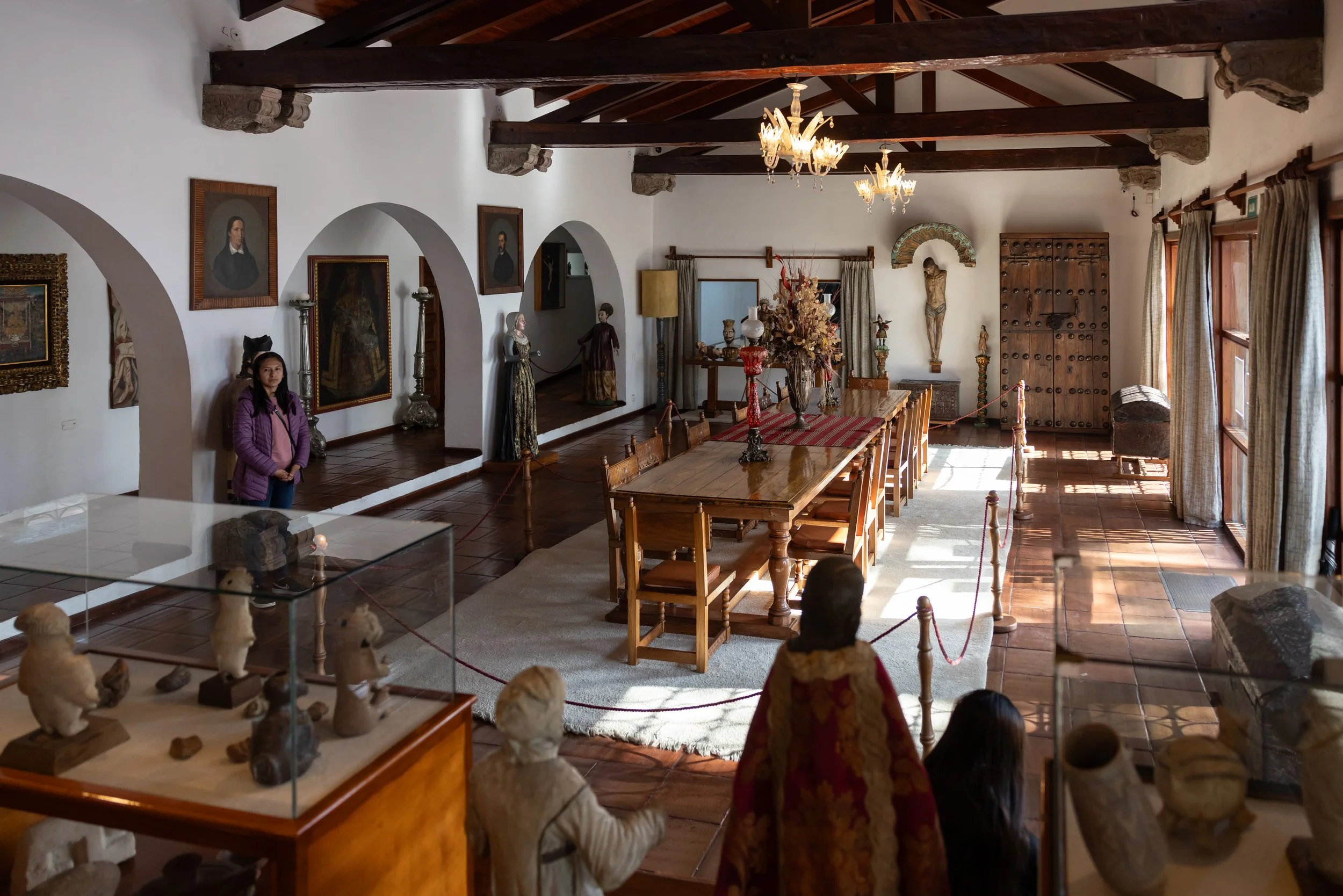 A spacious room with white walls and a wooden ceiling with beams, decorated with chandeliers, paintings, and statues. There are tables with vases and floral arrangements, along with various sculptures on display under a glass case. A woman stands nea